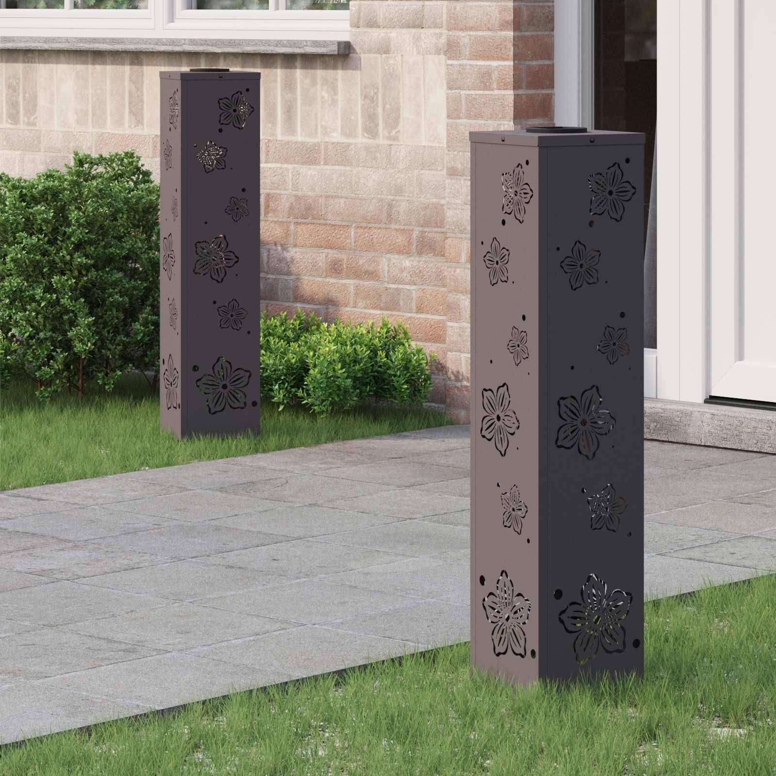 Dekorative Stehsäulen mit Blumenmustern an einem Gartenweg neben einem Ziegelgebäude und einer Tür, umgeben von Grünpflanzen.