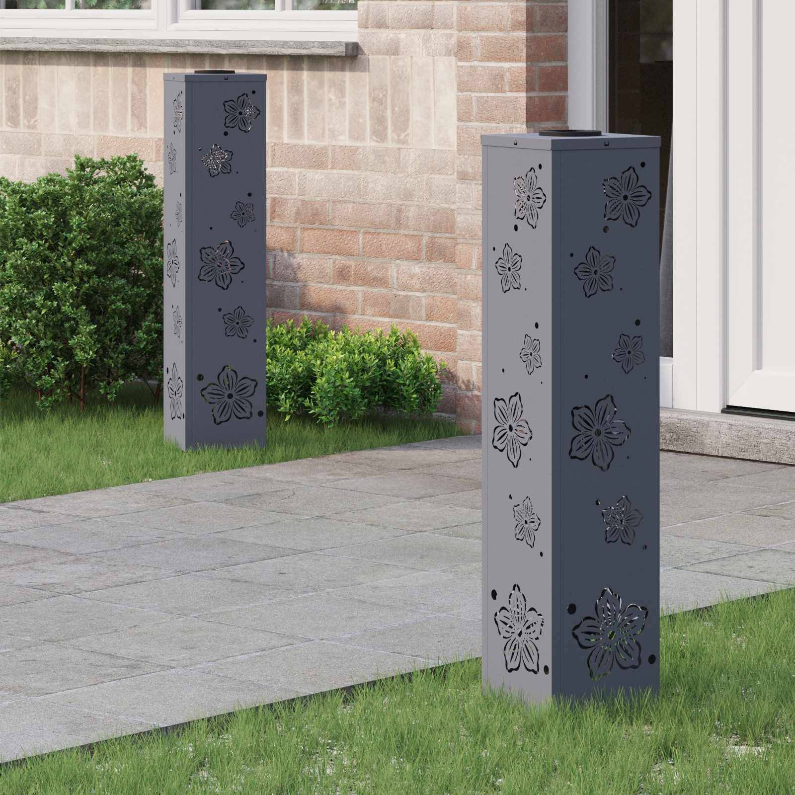 Dekorative Metallgartenpfähle mit Blumenmustern stehen auf einem Steinweg in der Nähe eines Backsteinhauses mit grünen Büschen und Gras.