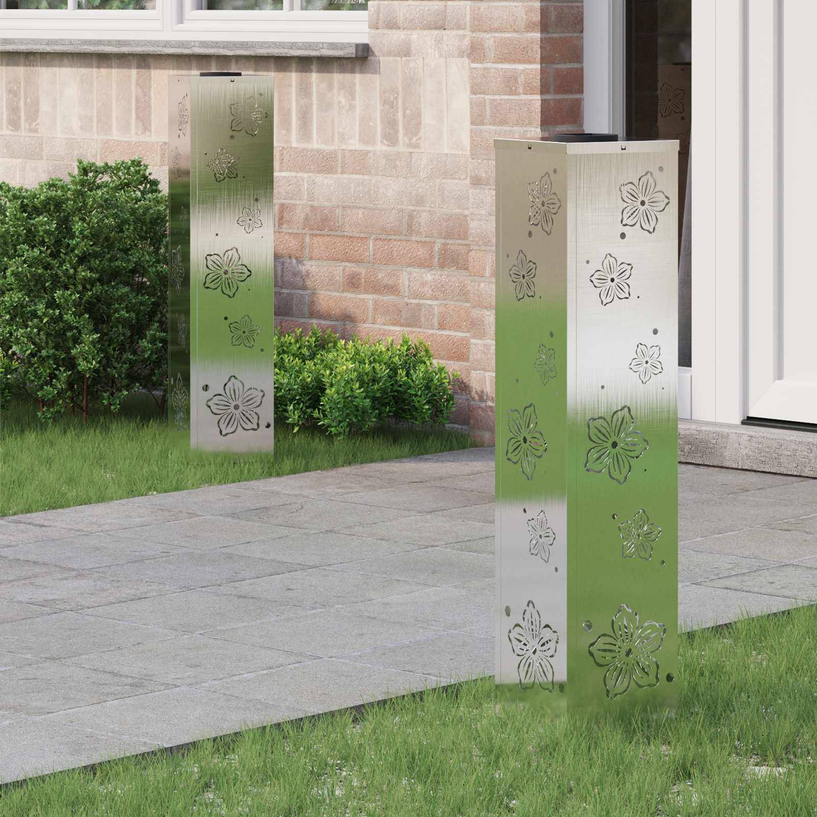 Zwei metallische Gartenleuchten mit Blumengravuren stehen auf einer Terrasse, umgeben von Gras und einer Backsteinmauer und verleihen einen modernen ästhetischen Eindruck.