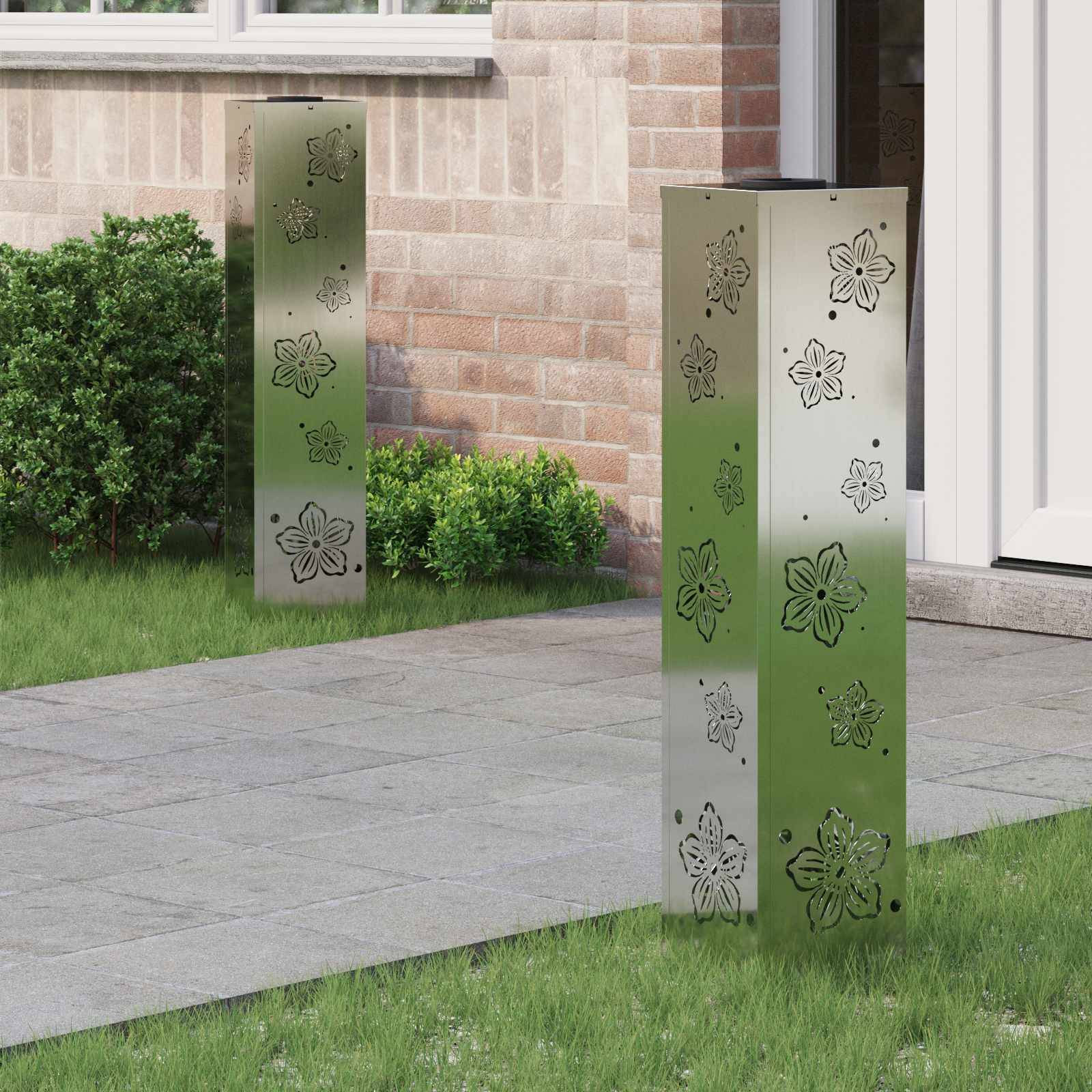 Zwei metallische Außenpfadleuchten mit Blumendurchbruchmustern sind auf einem Steinweg neben einer Backsteinmauer und grünen Sträuchern positioniert.