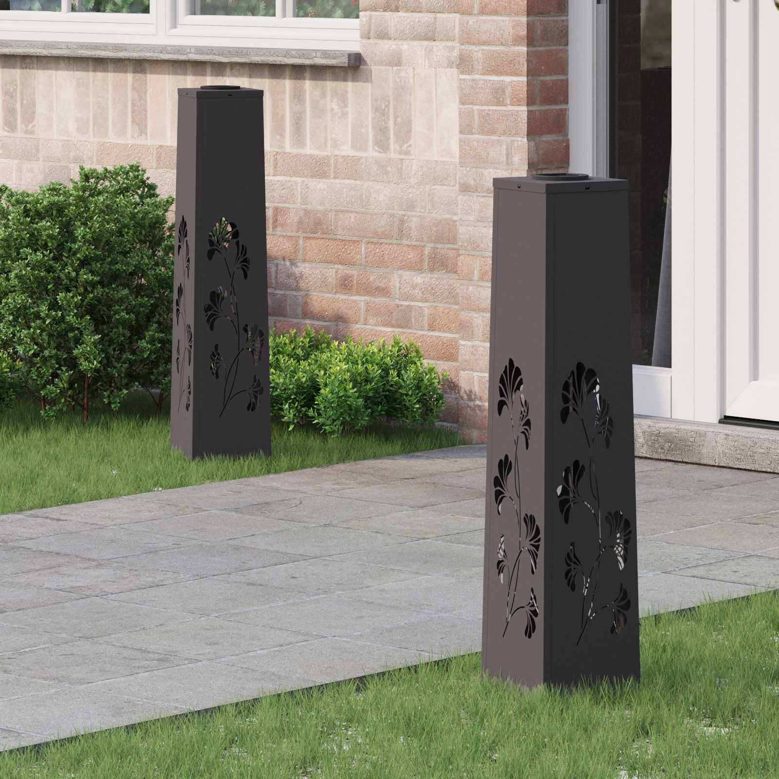 Zwei hohe, rechteckige Gartenlampen mit eingravierten Blattmustern stehen auf einem gepflasterten Weg, umgeben von Gras und einer Ziegelwand.