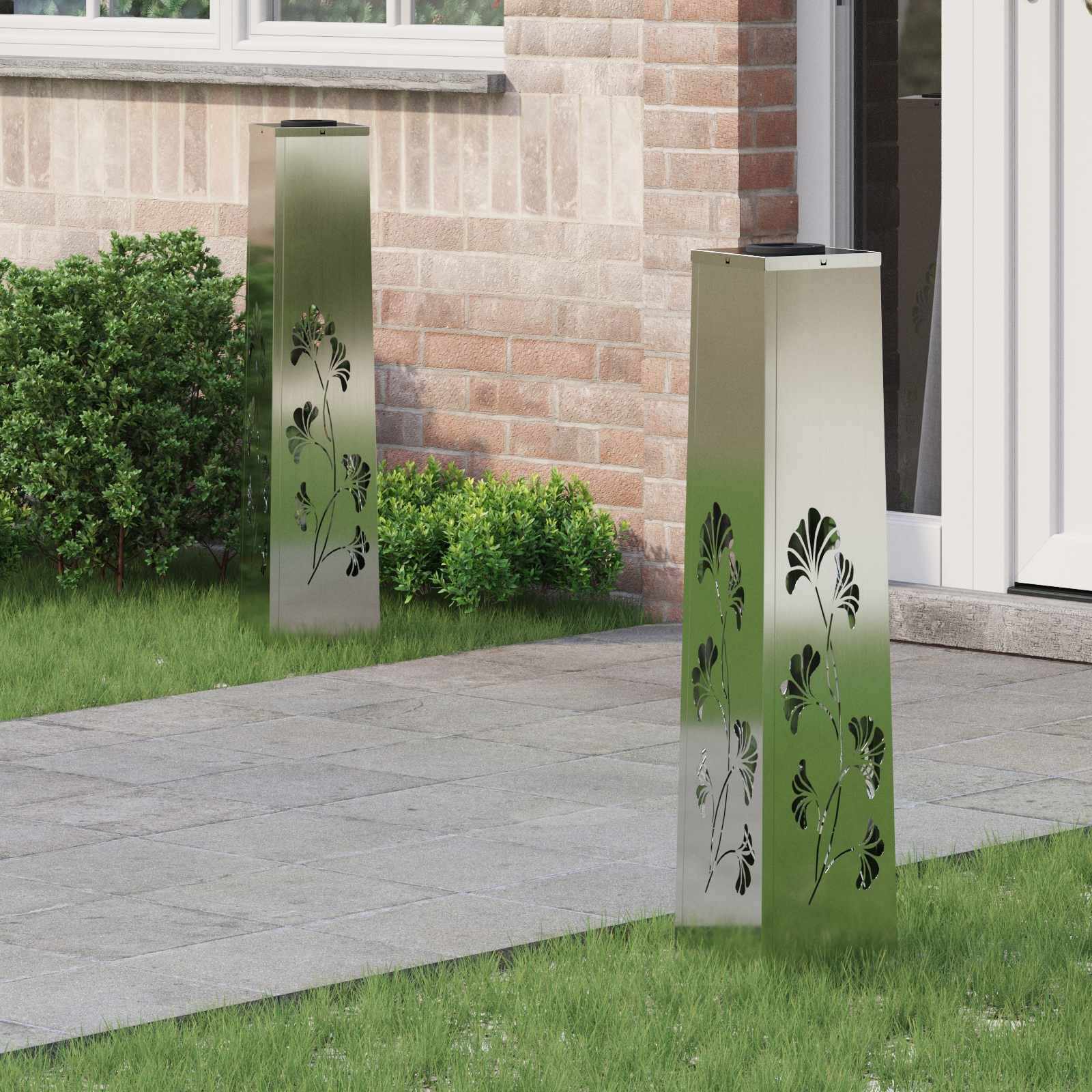 Außenszene mit zwei hohen, silbernen Außenlampenmasten mit Blattausschnitt-Designs, die auf einem Steinweg neben einer roten Ziegelmauer und grünen Sträuchern platziert sind.