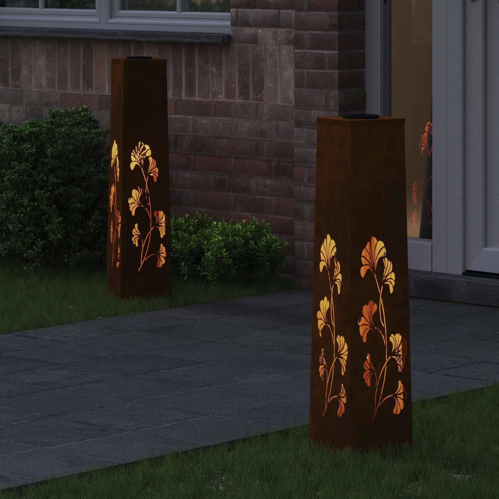 Metalllaternen mit Blumenmotiven aus Ausschnitten verbreiten warmes Licht entlang eines gepflasterten Gartenweges neben einer Ziegelwand und Gebüsch.