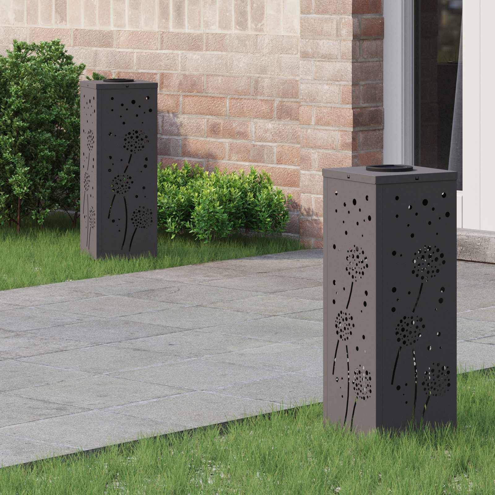 Zwei hohe, schwarze Außenlampen mit Löwenzahn-Ausschnitten stehen auf einem Steinweg in der Nähe von Sträuchern und einer Ziegelmauer und sorgen für dekorative Beleuchtung.