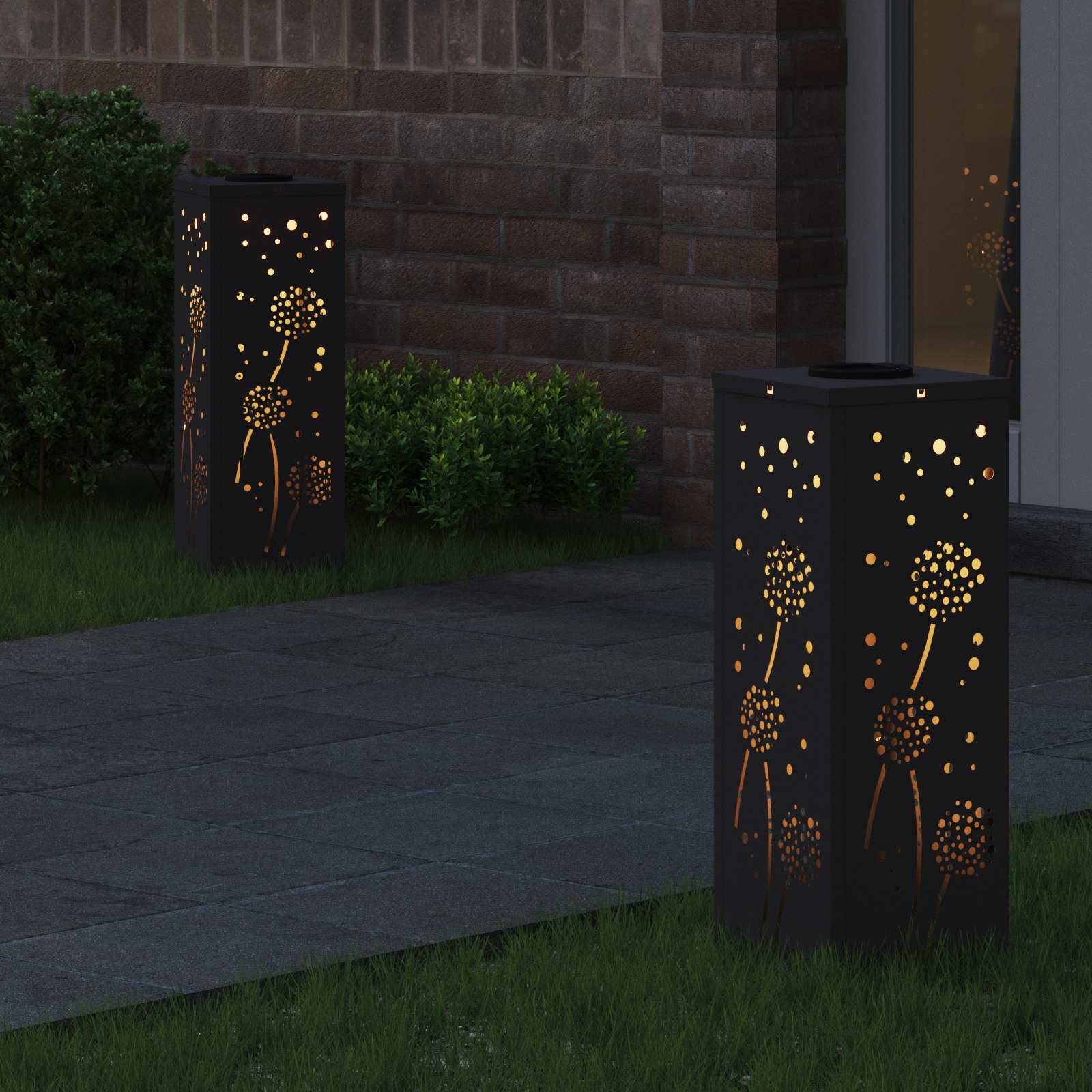 Zwei schwarze Laternen mit Löwenzahn-Ausschnitten leuchten sanft vor einem Eingang und beleuchten einen Steinweg, der von Gras und Sträuchern umgeben ist.