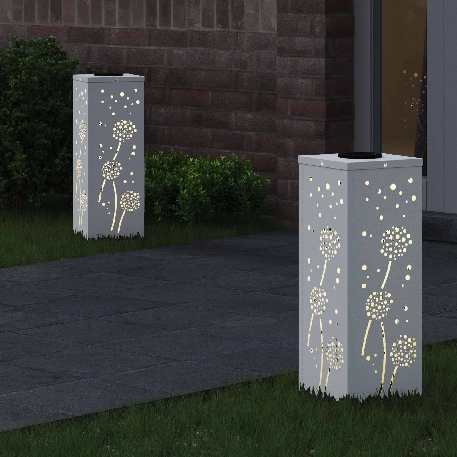 Zwei hohe, weiße Laternen mit Löwenzahn-Ausschnitten, die auf einem Rasen in der Nähe einer Ziegelwand platziert sind, verbreiten ein sanftes Leuchten und erzeugen eine beruhigende Atmosphäre.