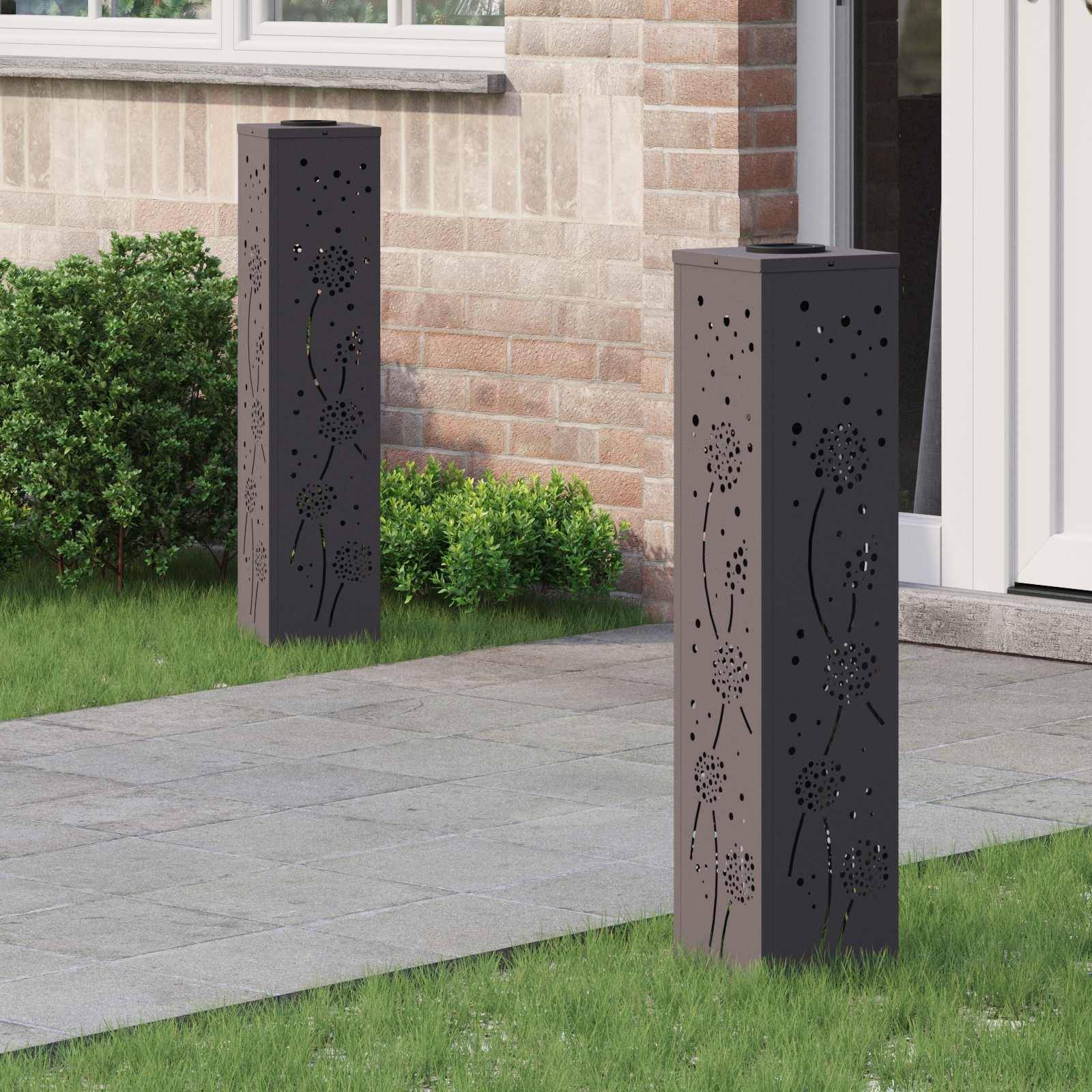 Zwei dekorative Metallpfosten mit floralem Durchbruchmuster stehen neben einem Weg in der Nähe einer Ziegelwand und grüner Strauchvegetation.