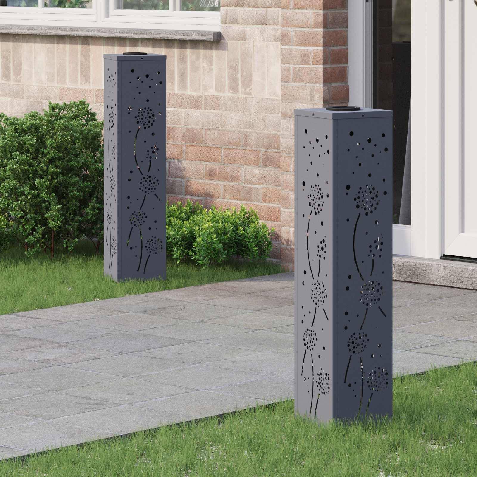 Zwei dekorative Metallsäulen mit Löwenzahn-Ausschnitten stehen auf einem gepflasterten Weg vor einem Backsteinhaus. Grüne Sträucher säumen den Pfad.