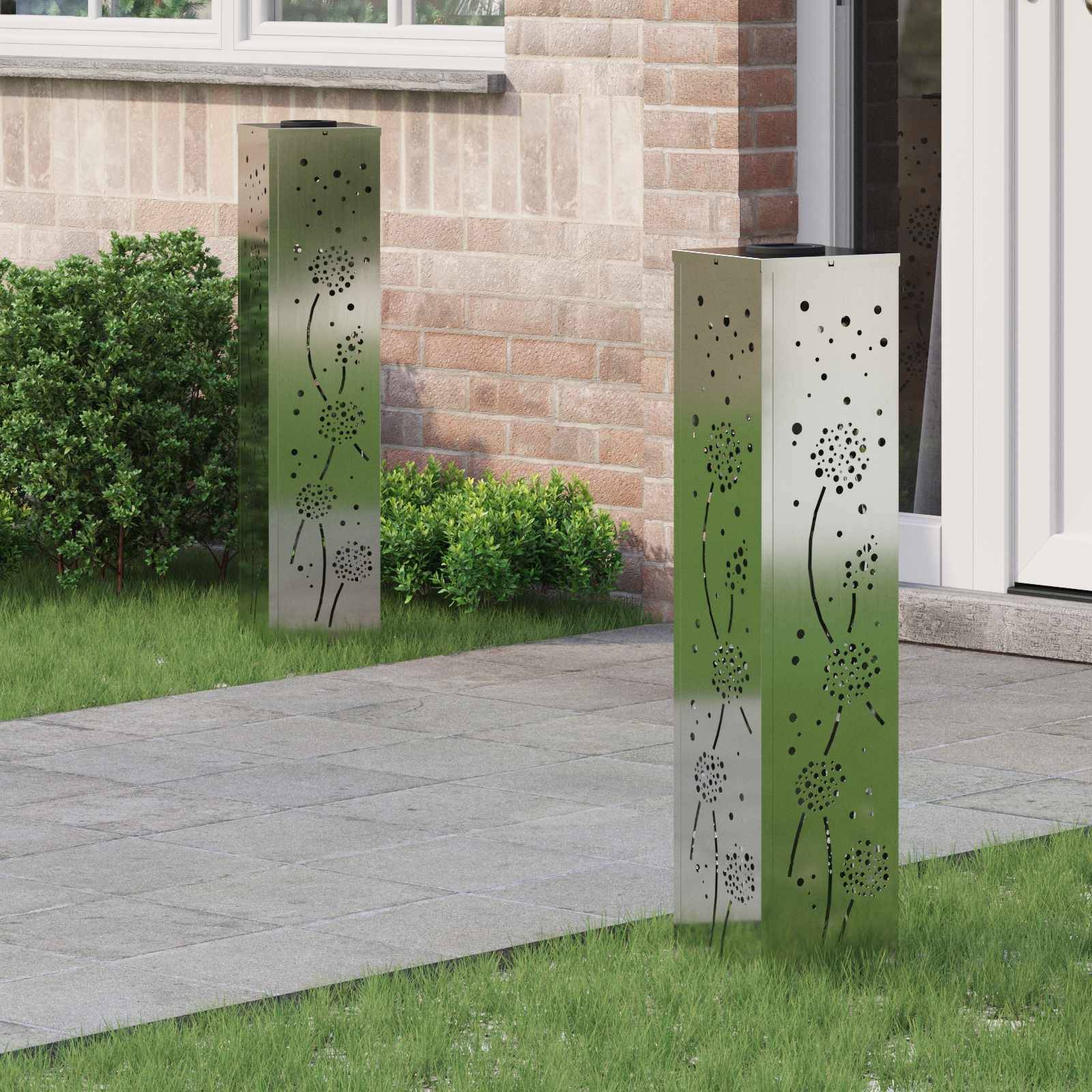 Zwei dekorative Außenlampen mit floralen Ausschnitten stehen auf einem gepflasterten Weg neben einem Backsteinhaus, umgeben von grünen Büschen und Gras.