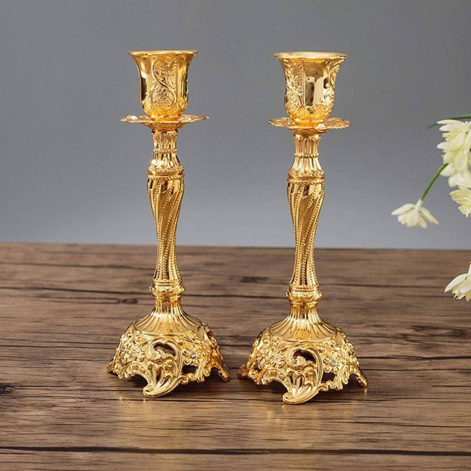 Zwei verzierte Goldkerzenhalter mit kunstvollen Designs stehen auf einer Holzoberfläche. Eine weiße Blume ist teilweise rechts sichtbar.