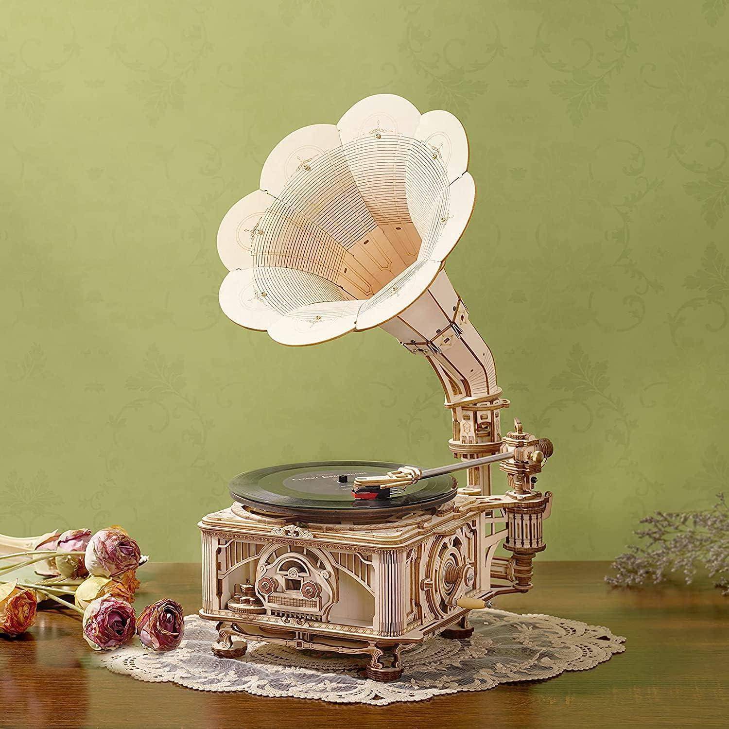 Ein Grammophon im Vintage-Stil mit einer blumenförmigen Trichtermuschel steht auf einem mit einer Deckchen bedeckten Tisch, umgeben von verstreuten Blumen vor einem grünen Hintergrund.