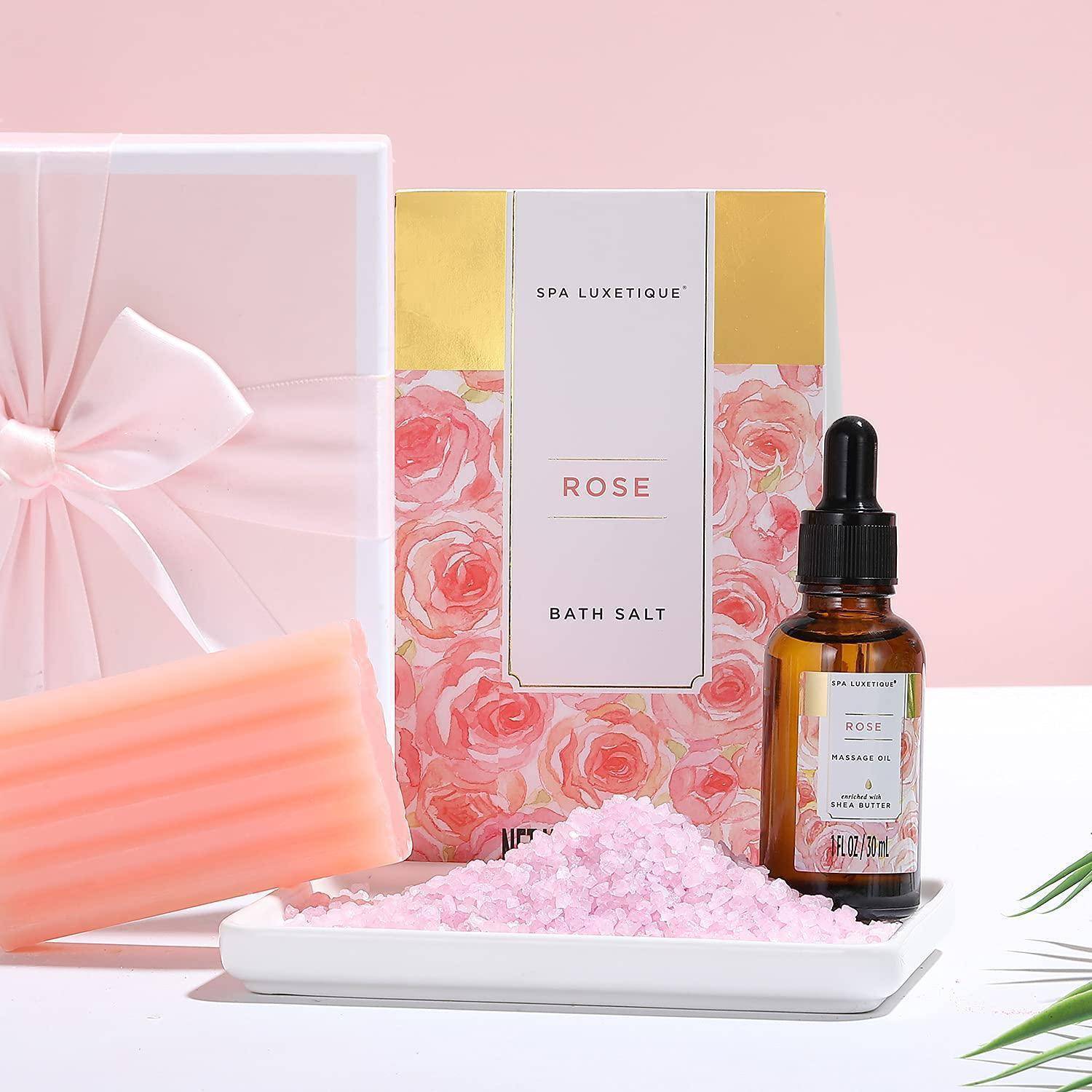 Geschenkset 'Rosen-Badesalz' mit einer rosa Seifenbar, rosa Salz und einer braunen Flasche auf einer weißen Oberfläche, mit einer rosa und goldenen Box im Hintergrund.