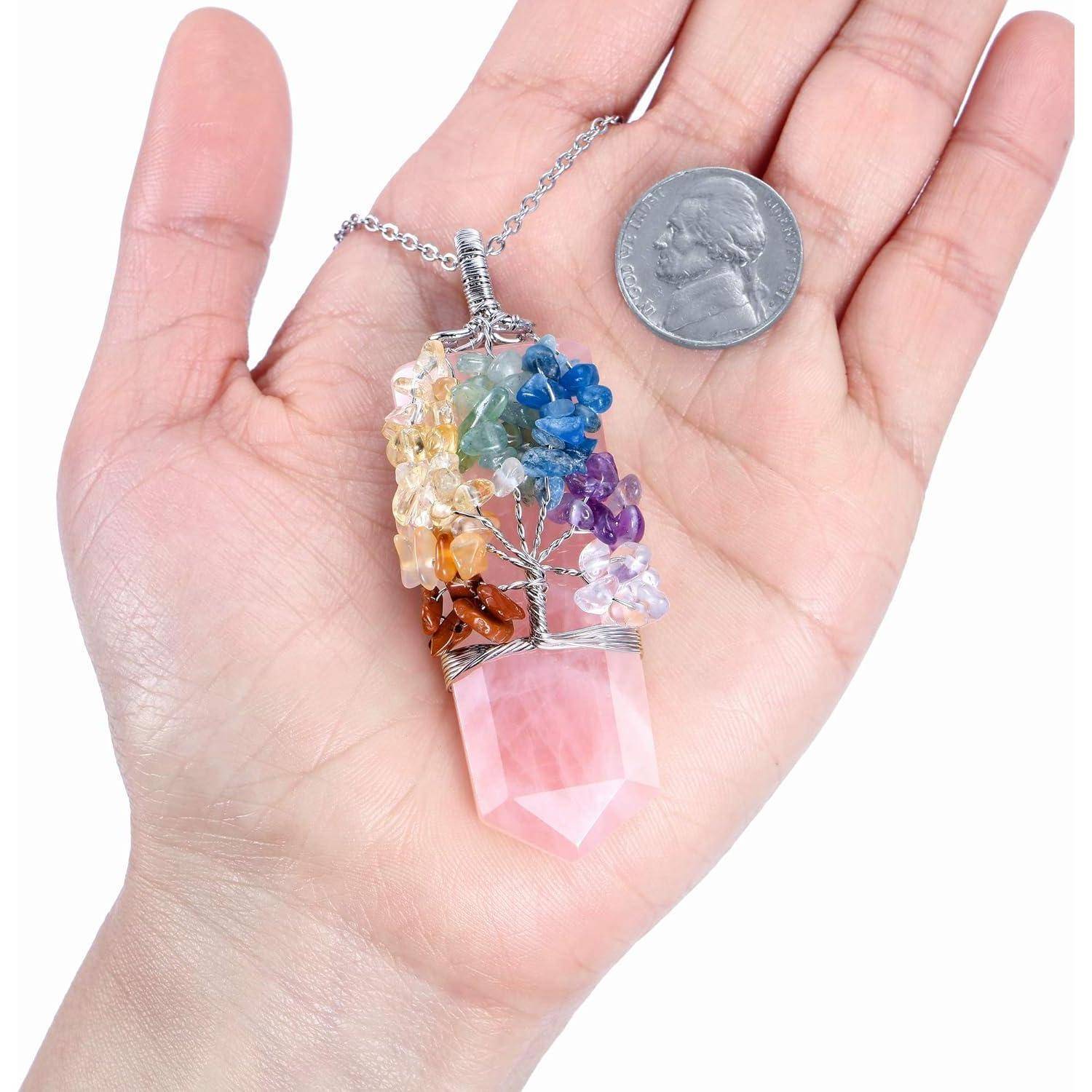 Eine Hand hält eine silberne Halskette mit einem rosa Kristallanhänger und einem bunten Edelsteinbaum, der darüber mit Draht umwickelt ist, neben einer Münze zur Größenbestimmung.