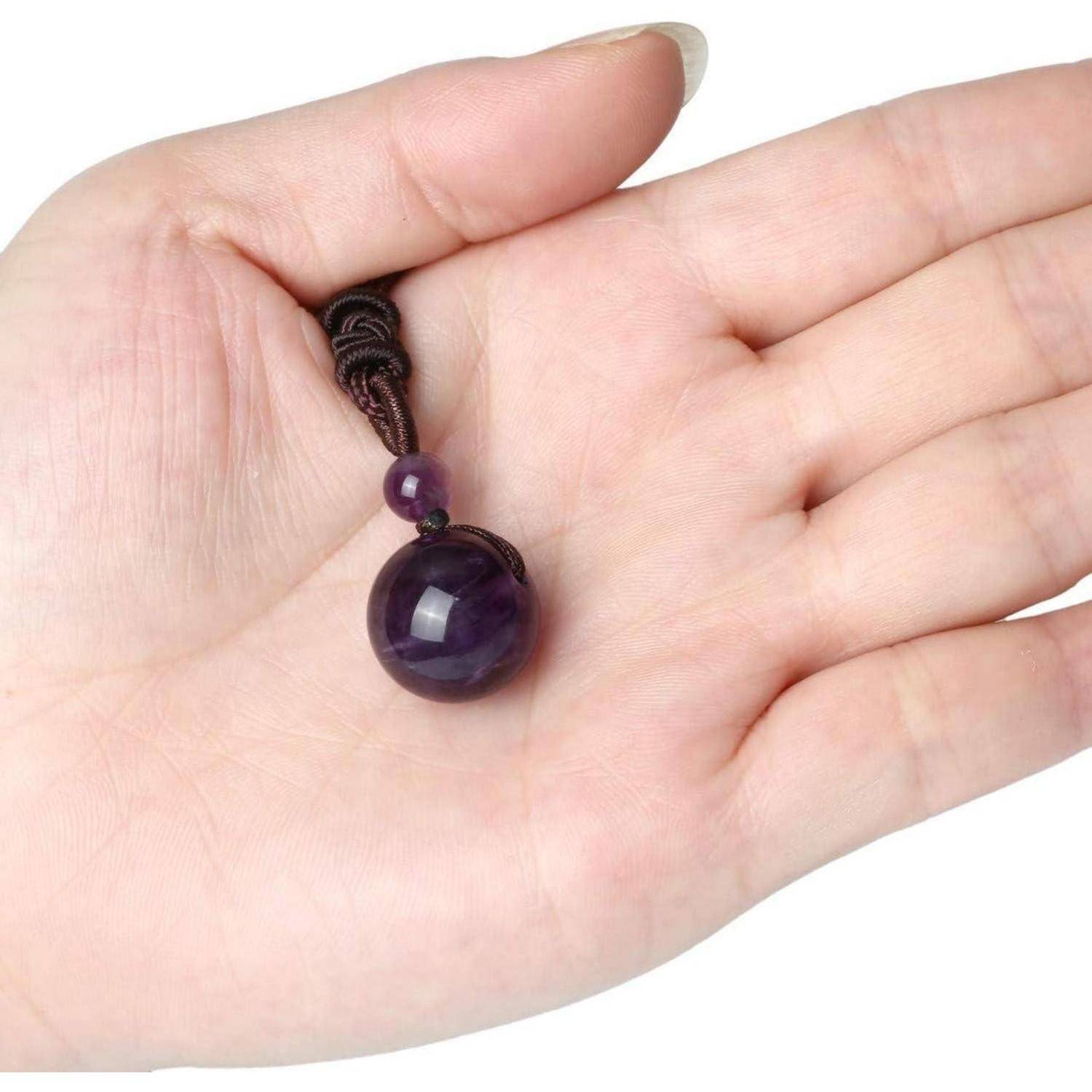 Eine Hand, die eine lila sphärische Perle mit einer kleinen, benachbarten Perle hält, beide an einer braunen Schnur mit einem Knoten befestigt, vor einem einfarbigen Hintergrund.
