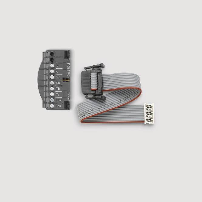 Ein schwarzer Stecker mit beschrifteten Pins ('a', 'b', 'c', 'GND', 'TxD', 'RxD', 'TaK'), der an ein flaches, mehrfarbiges Flachbandkabel auf grauem Hintergrund angeschlossen ist.