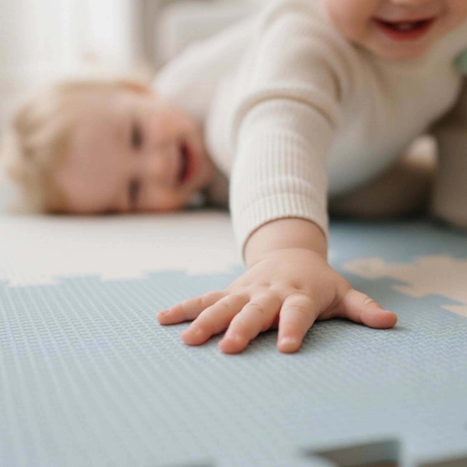 Ein kleines Kind kriecht auf einer blauen Schaumstoff-Spielmatte und streckt eine Hand aus, während ein anderes Kind im Hintergrund liegt und lächelt.