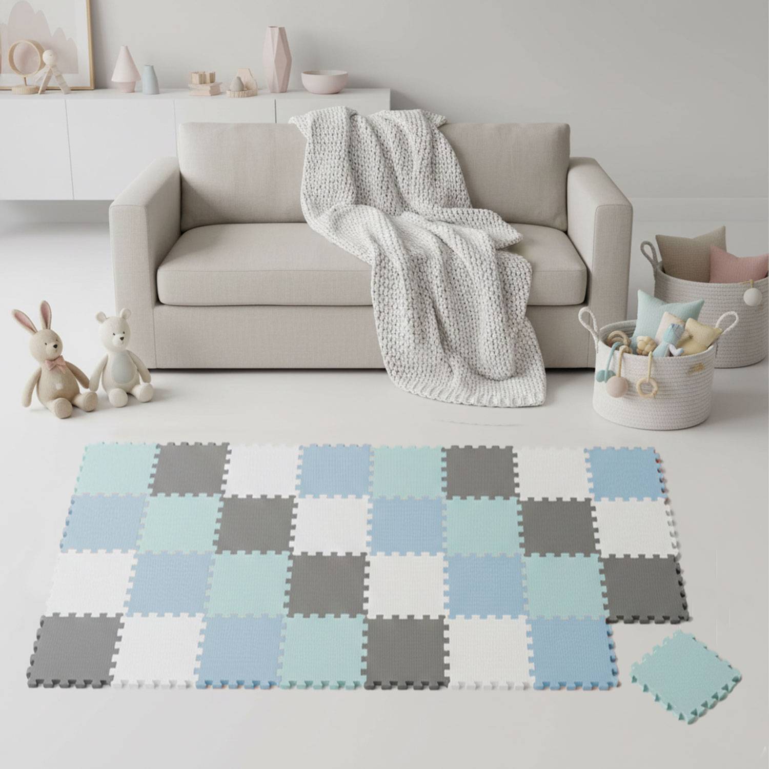 Ein neutral gehaltener Wohnbereich zeigt ein hellgraues Sofa mit einer weißen gestrickten Decke. Zwei Plüschtiere sitzen auf einer Schachbrettmuster-Spielmatte.