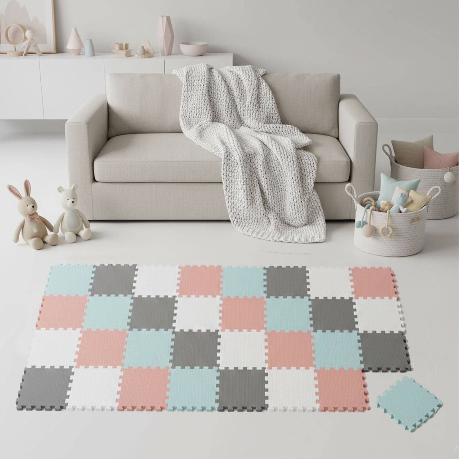 Ein pastellfarbener Spielraum zeigt ein hellgraues Sofa mit einer gestrickten Decke, zwei Plüschhasen und einer Patchwork-Schaumstoff-Spielmatte auf dem Boden.