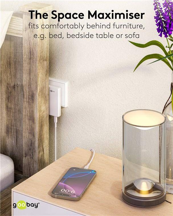 Ein Smartphone mit sichtbarem Ladebatteriesymbol liegt auf einem Holznachttisch neben einer Glühlampe, einem Pflanzenstängel und einer Wandsteckdose.