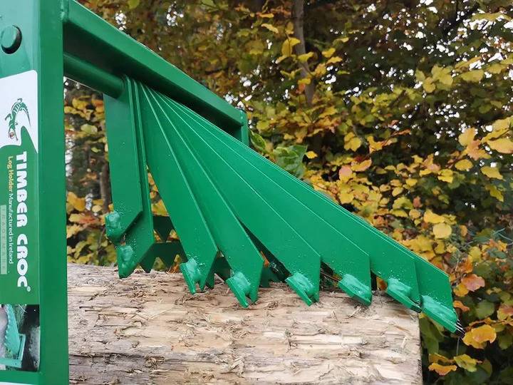 Ein grüner Metallholzspalter, der auf einem Baumstamm positioniert ist, mit scharfen Keilen, die bereitstehen, den Holzblock zu spalten, vor einem Hintergrund von herbstlichem Laub.