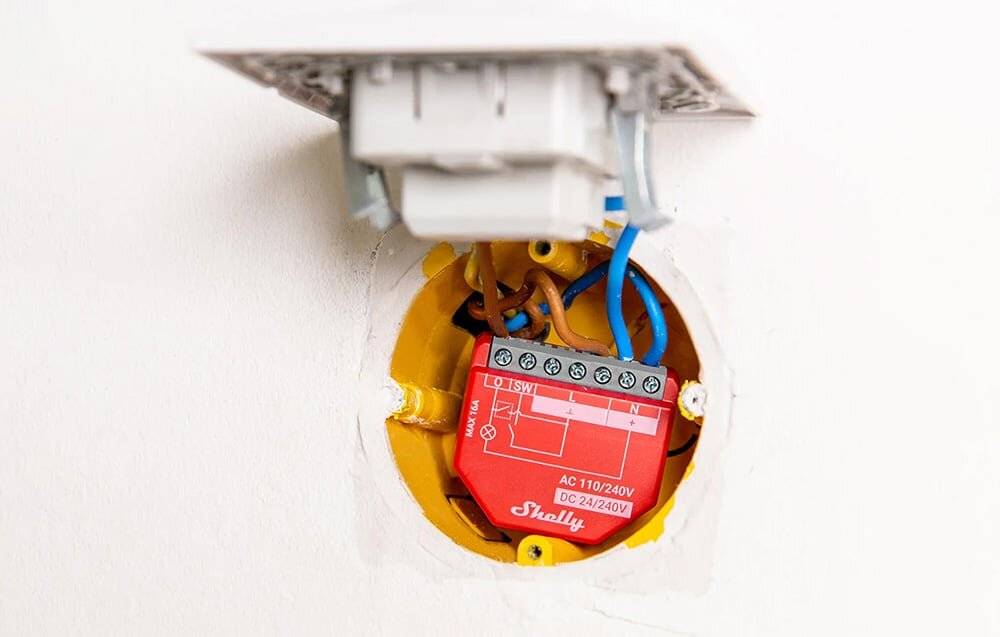 Elektrische Einrichtung mit einem roten Shelly-Relais, das mit Drähten in einer Wandsteckdose verbunden ist. Die Konfiguration zeigt AC 110/240V und DC 24/240V.