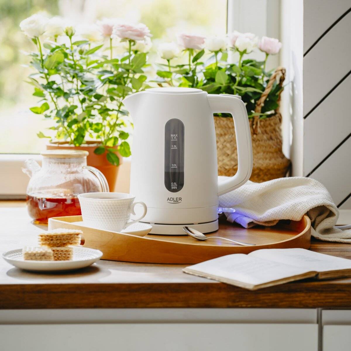 Elektrischer Wasserkocher auf der Küchenablage mit Tasse, Untertasse und Zuckerwürfeln. Heller Fensterblick mit Pflanzen draußen und einem offenen Notizbuch in der Nähe.