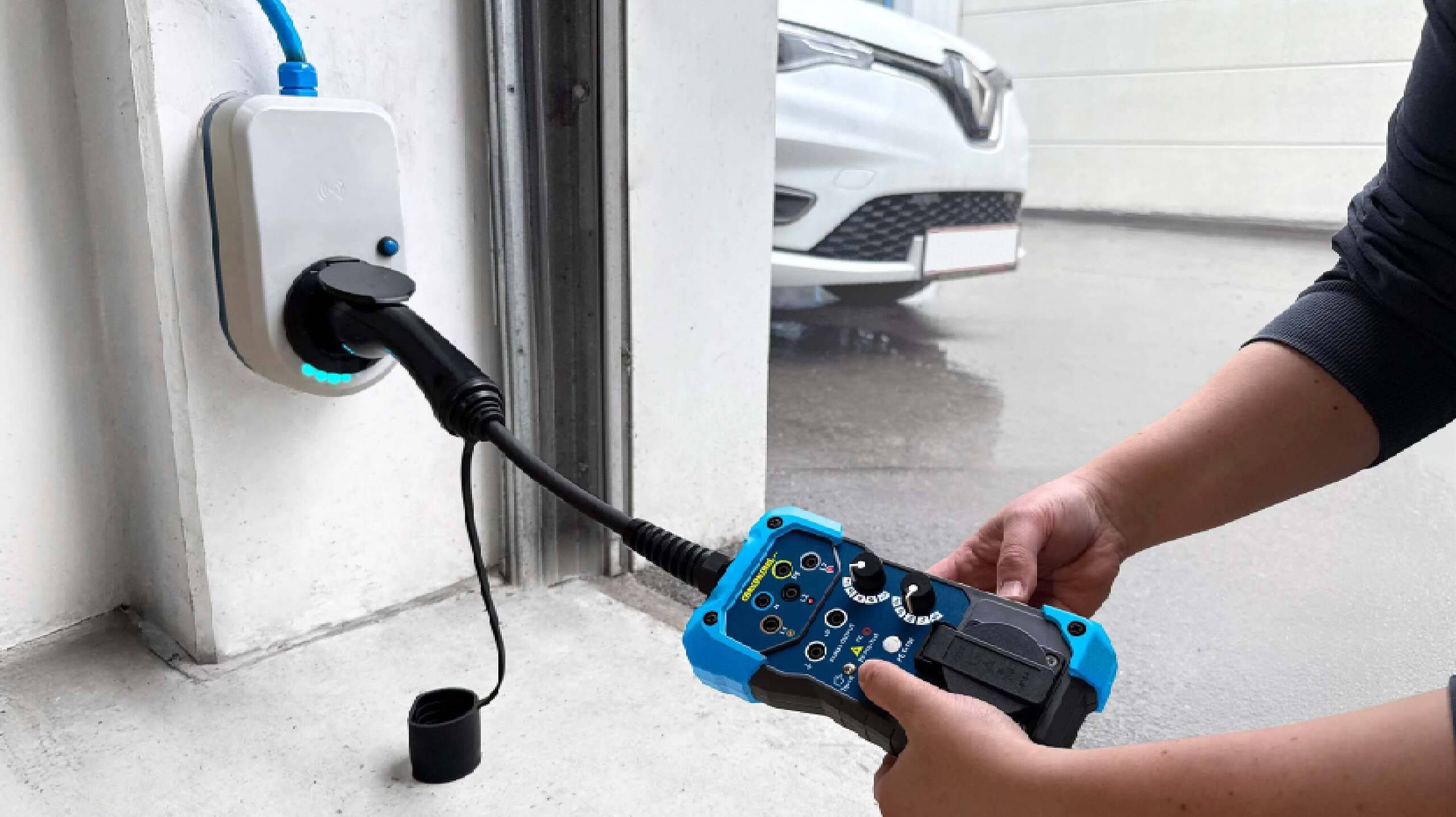 Eine Hand hält eine Ladekontrolleinheit für Elektrofahrzeuge, wobei das Ladegerät in eine Wandsteckdose eingesteckt ist. Ein Auto ist in der Nähe geparkt.