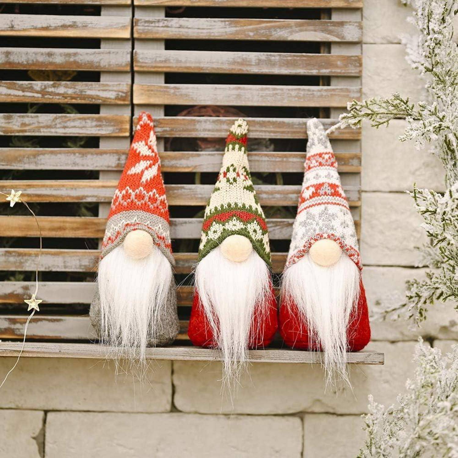 Drei gestrickte Gnome mit langen weißen Bärten und bunten Mützen sitzen auf einem Holzregal vor einer Ziegelwand, umgeben von winterlicher Dekoration.