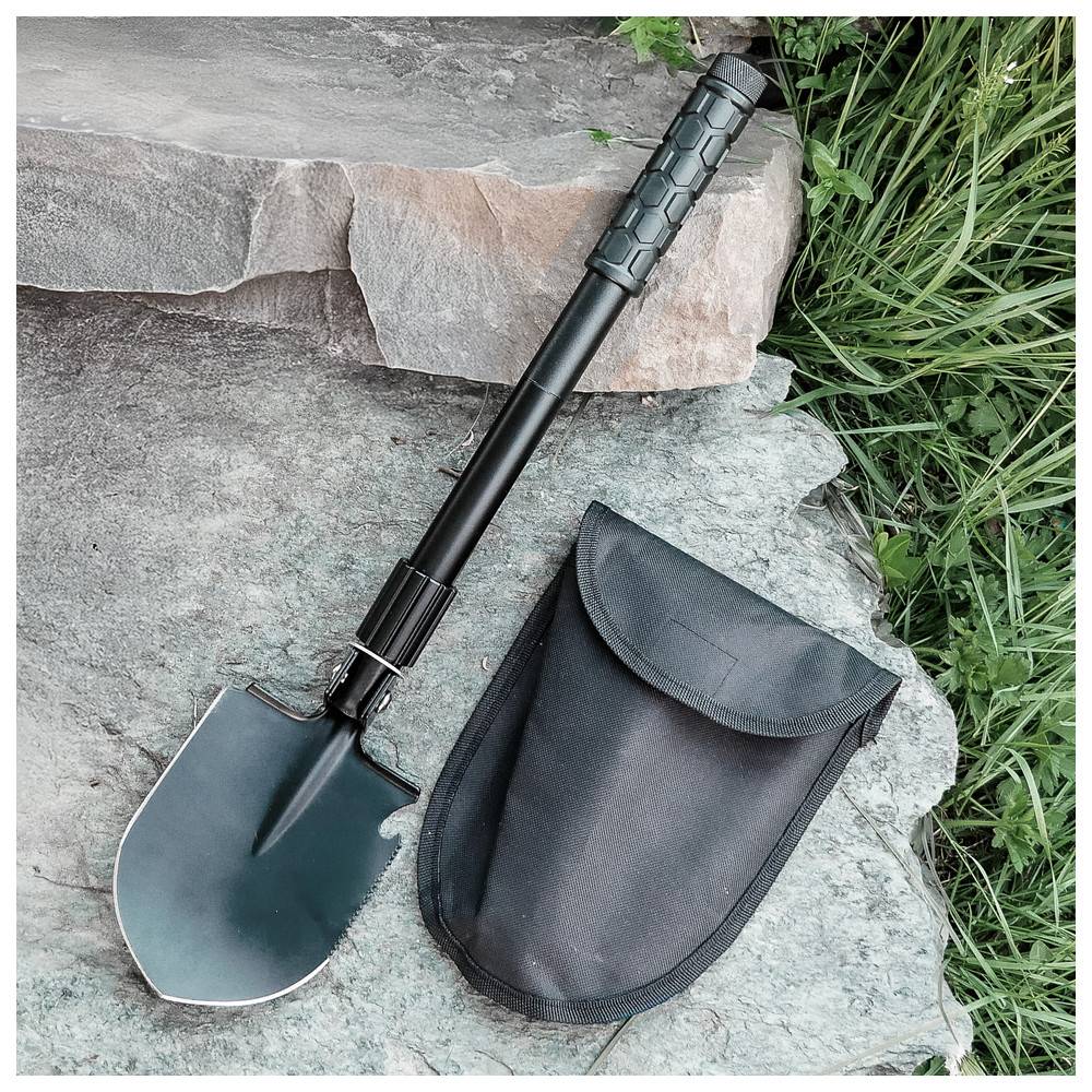 Eine zusammenklappbare Schaufel mit einem schwarzen Griff liegt neben ihrer schwarzen Transporttasche auf einer Steinoberfläche, umgeben von grünem Gras.