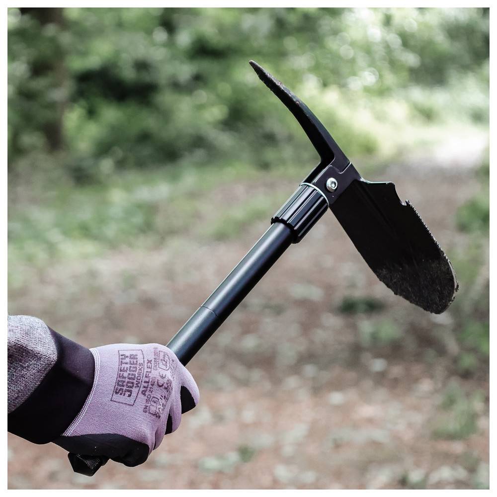 Eine Person mit Handschuhen hält eine kleine schwarze Spitzhacke über einem Erdweg in einem bewaldeten Gebiet und deutet damit auf Außenarbeit oder Gartenarbeit hin.