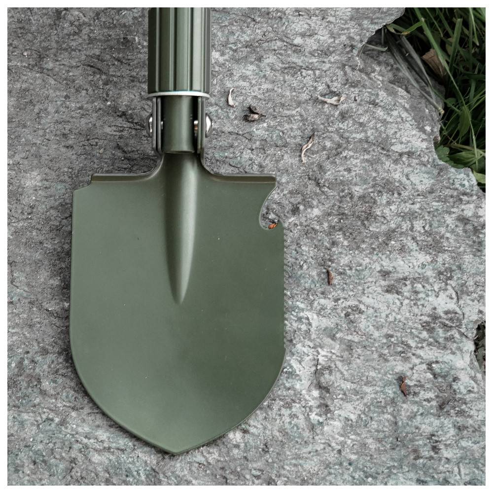 Ein grüner, metallener Schaufelkopf liegt auf einer rauen, grauen Steinoberfläche, mit einigen kleinen Zweigen in der Nähe verstreut.