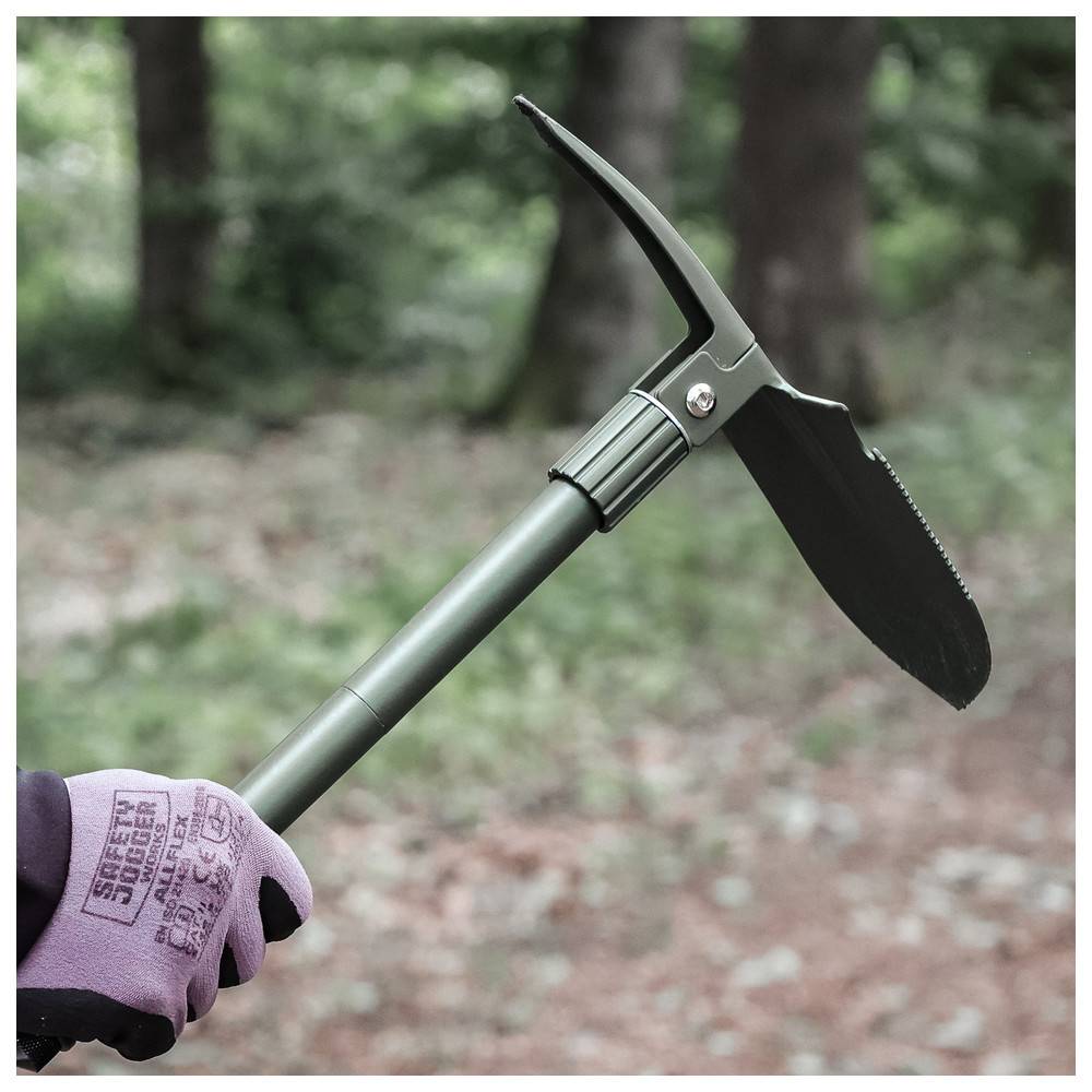 Eine Person, die violette Handschuhe trägt, hält einen militärischen Klappspaten mit gezackter Kante in einer Waldlandschaft.