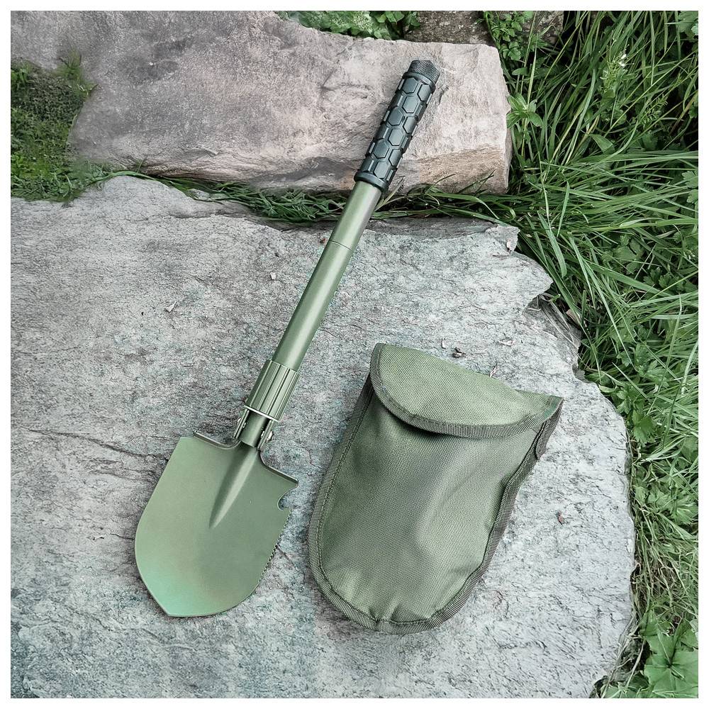 Eine Klappschaufel in Grün mit einem strukturierten Griff liegt auf einem Felsen neben einem grünen Stoffbeutel, umgeben von Gras und weiteren Steinen.