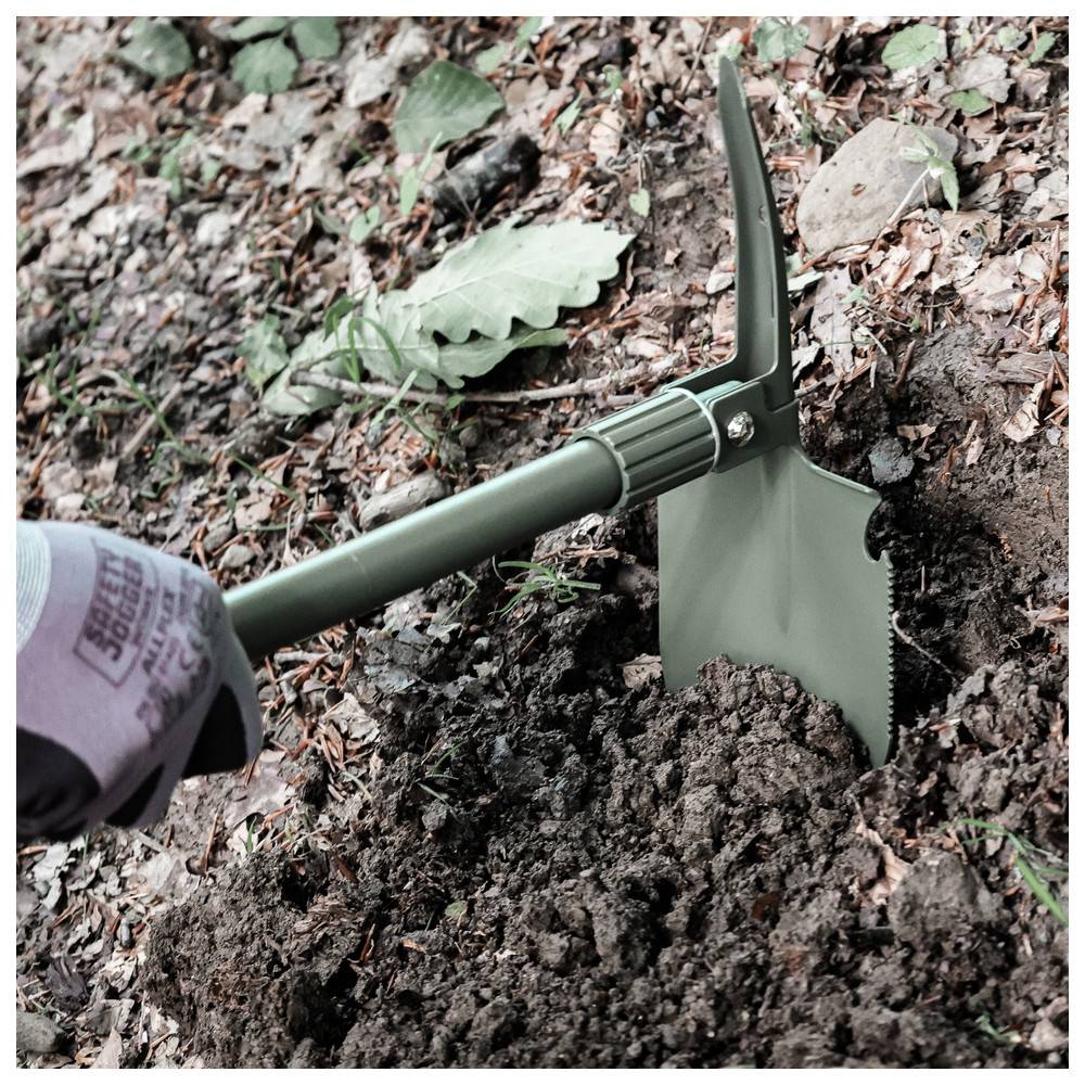 Eine Person benutzt einen grünen Klappspaten, um Erde in einem bewaldeten Gebiet auszuheben, wobei sichtbare Blätter und kleine Steine um die Grabstelle herum liegen.