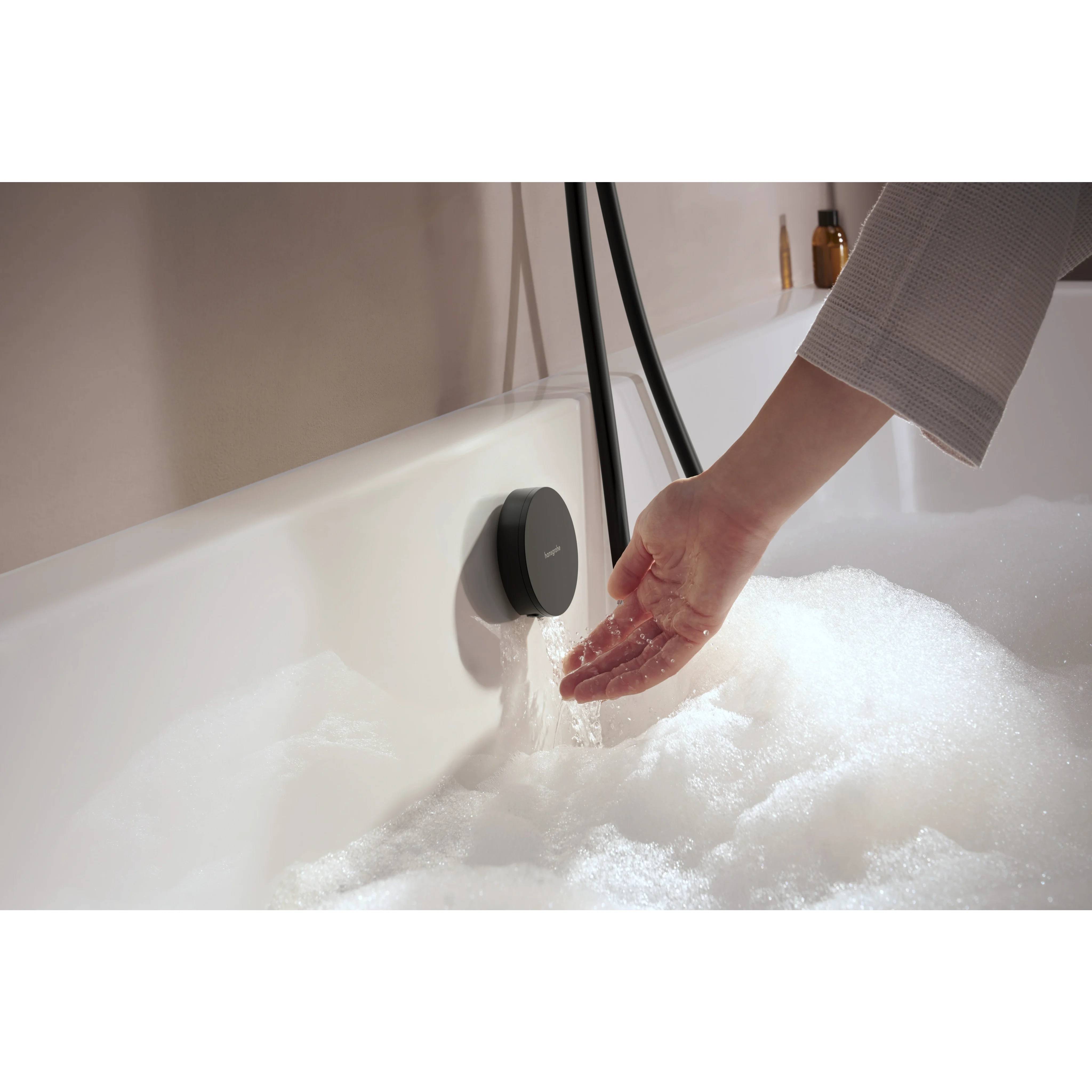 Eine Hand testet Wasser, das aus einem Badewannenhahn in eine mit Schaum gefüllte Wanne fließt.