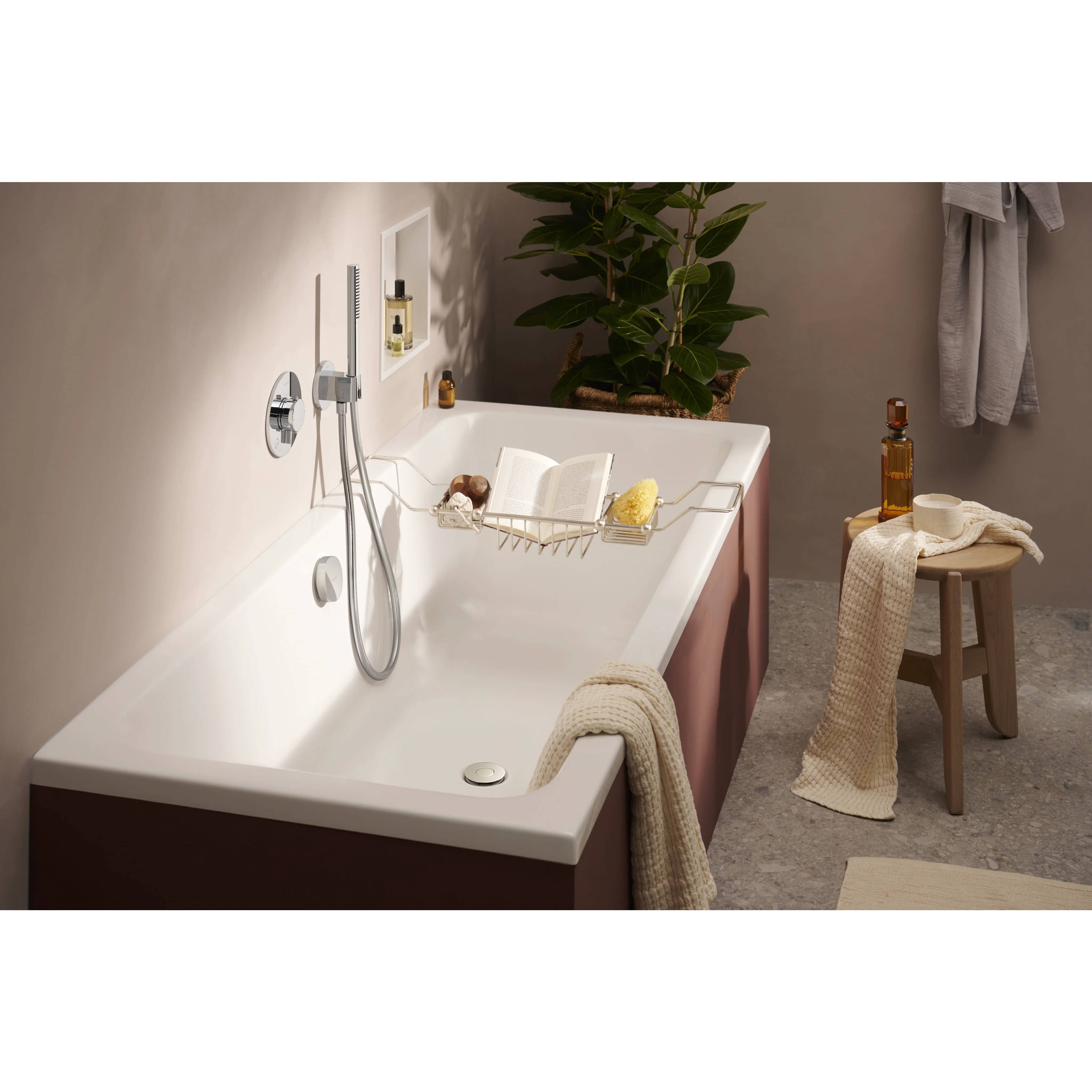 Modernes Badezimmer mit einer weißen Badewanne, einer wandmontierten Dusche, einem Badewannenablage, einem Hocker mit Handtuch und Flasche sowie einer Pflanze im Hintergrund.