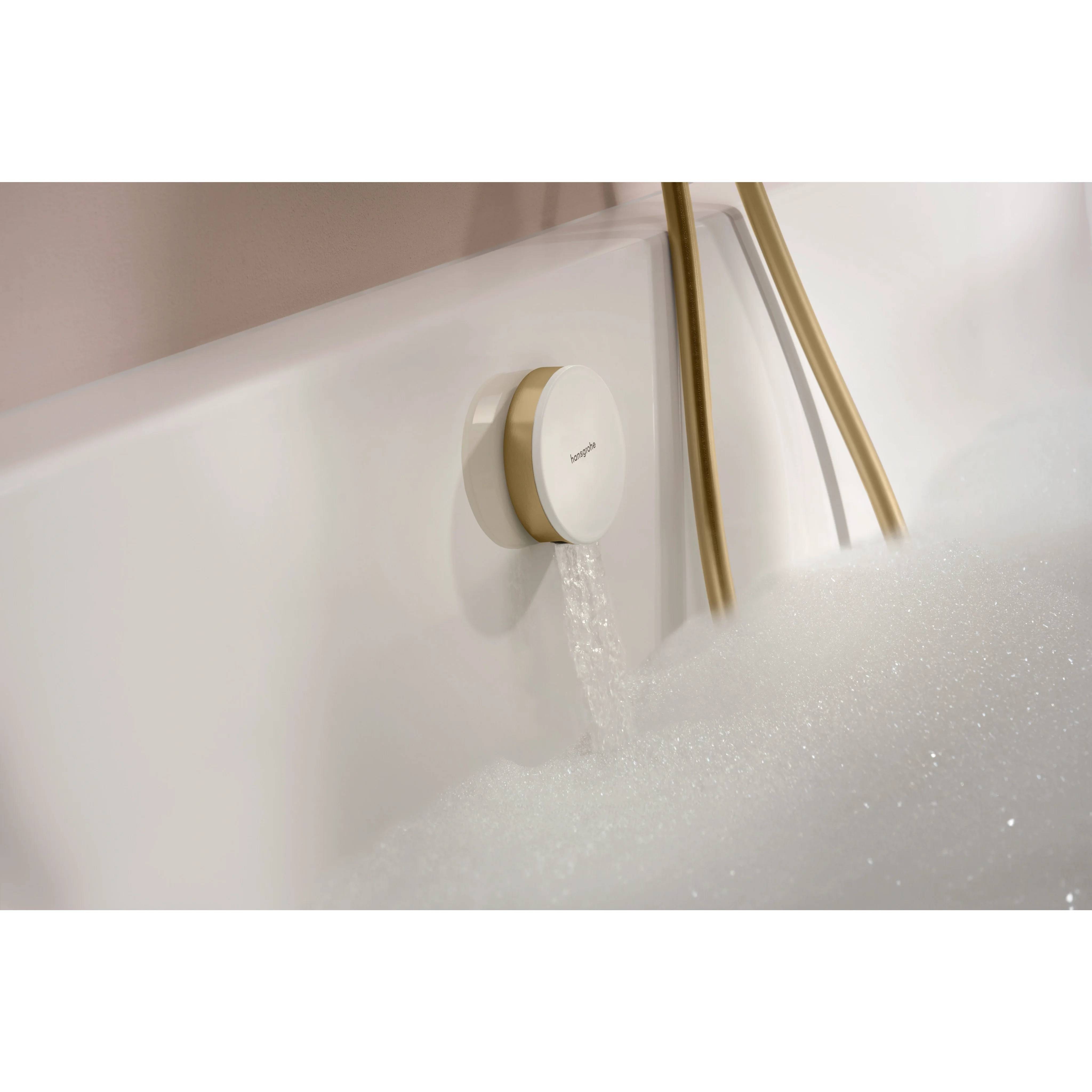 Wasser fließt aus einem runden Badewannenhahn in eine Wanne, die mit weißem Schaum gefüllt ist, wobei goldfarbene Brauseschläuche sichtbar sind.