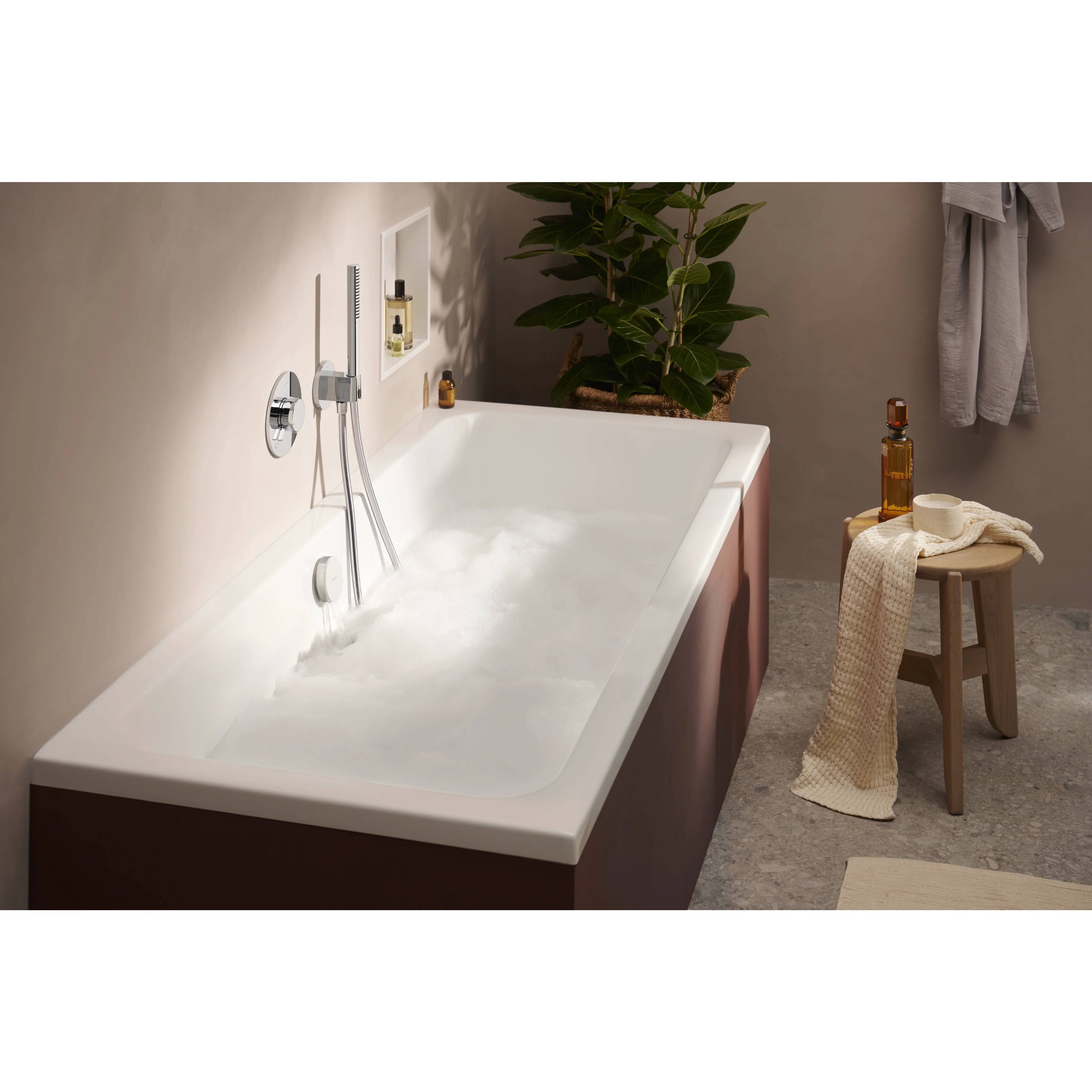 Freistehende Badewanne mit Schaum in einem modernen Badezimmer, mit einer Duscharmatur, einem Hocker, einem Handtuch und einer Pflanze in der Nähe.