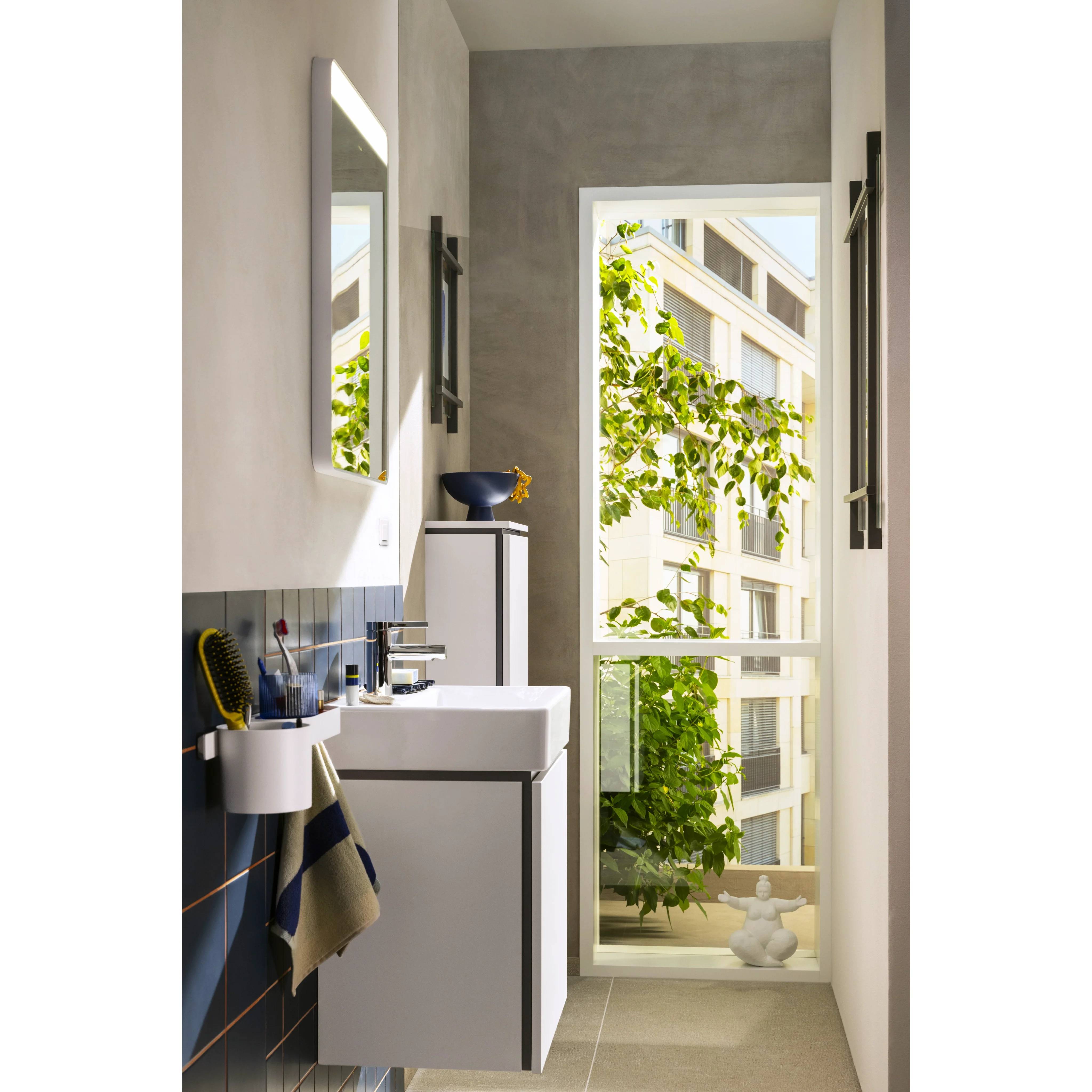 Modernes schmales Badezimmer mit an der Wand montiertem Waschbecken und Spiegel, mit Blick auf ein Fenster mit Aussicht auf grüne Bäume und ein nahegelegenes Apartmentgebäude.