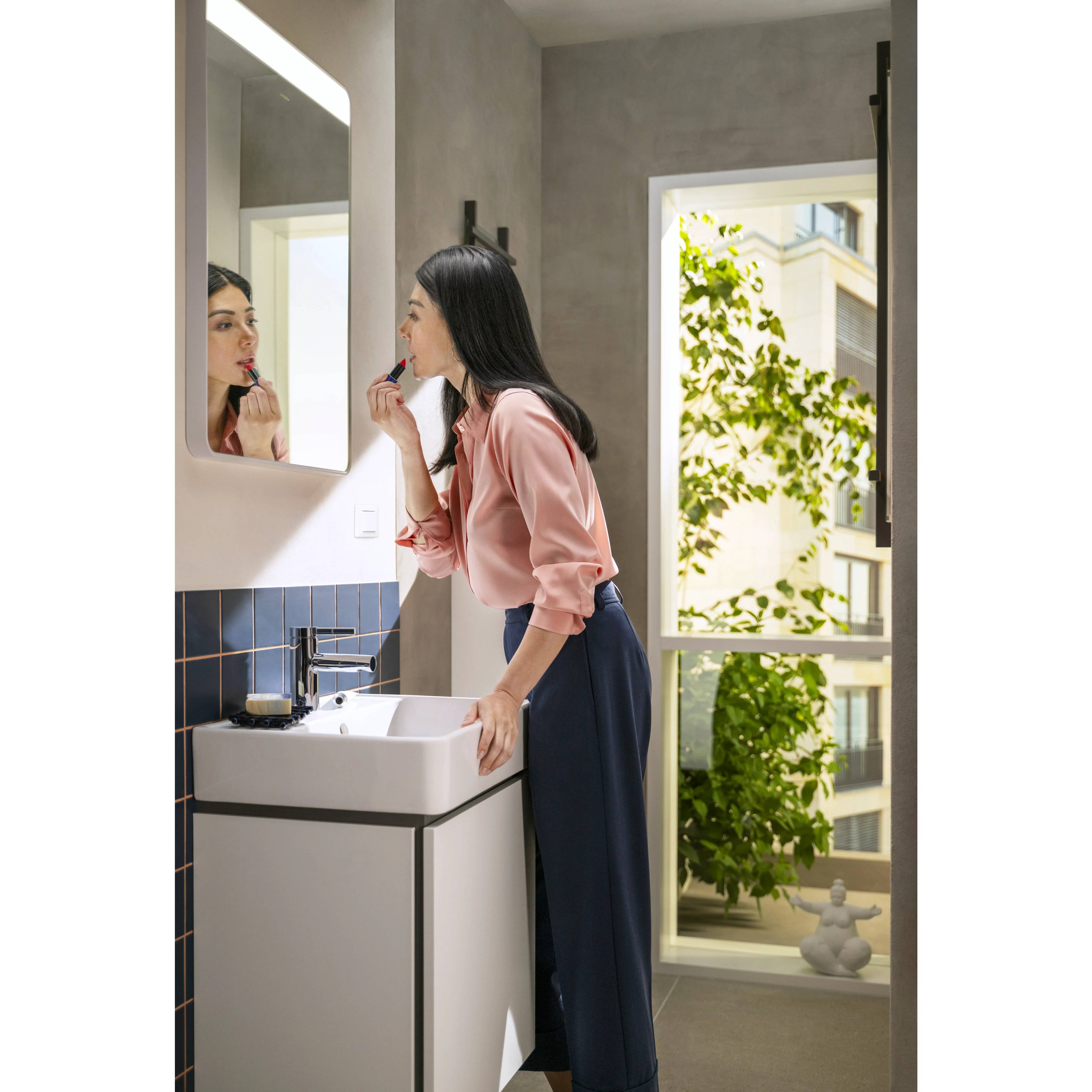 Frau trägt Lippenstift an einem Waschbecken im Badezimmer auf, während sie in den Spiegel schaut, mit einem offenen Fenster und Grünflächen draußen.