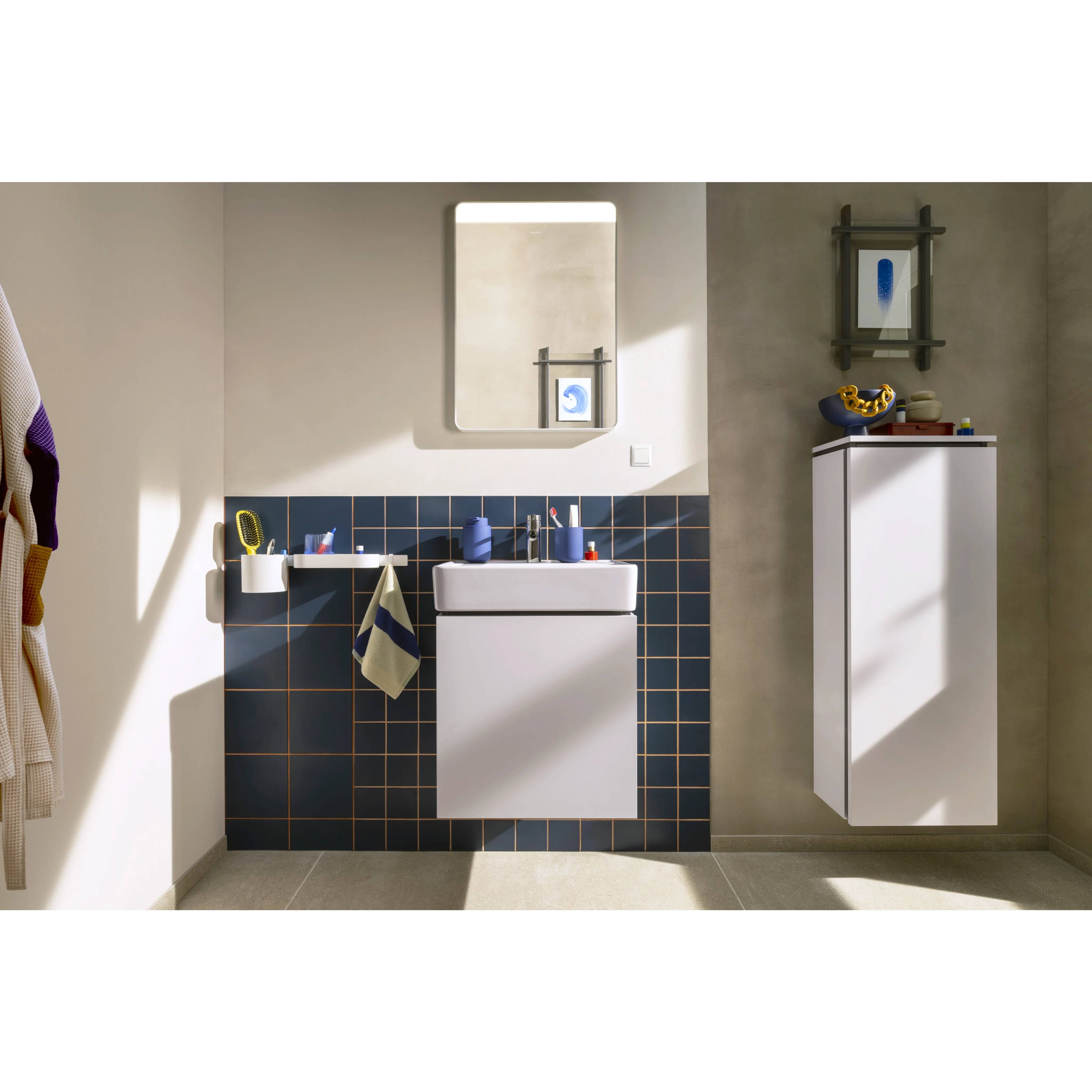 Modernes Badezimmer mit wandmontiertem Waschbecken, Spiegel, dunkelblauer Fliesenrückwand und hohem Schrank auf der rechten Seite bei hellem Sonnenlicht.