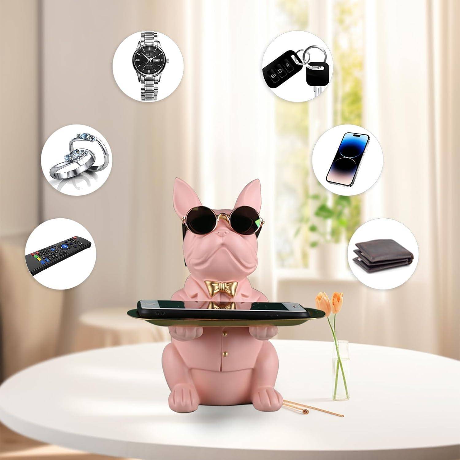 Eine Hundestatue in Rosa, die eine Sonnenbrille trägt und ein Tablett mit einem Telefon hält. Umgebende Gegenstände umfassen eine Uhr, Autoschlüssel, Telefon, Geldbörse, Ring und Fernbedienung.