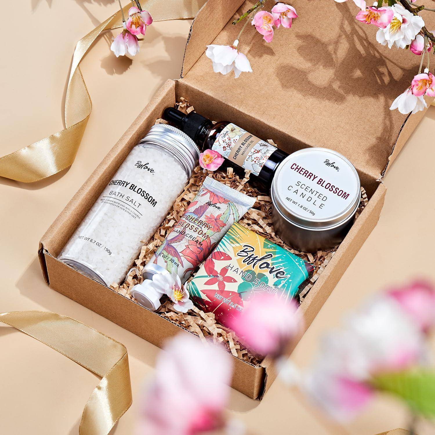 Eine Geschenkbox gefüllt mit kirschblütenthematischen Artikeln: Badesalz, Kerze, Handcreme, Seife und Raumspray, umgeben von rosa Blumen.