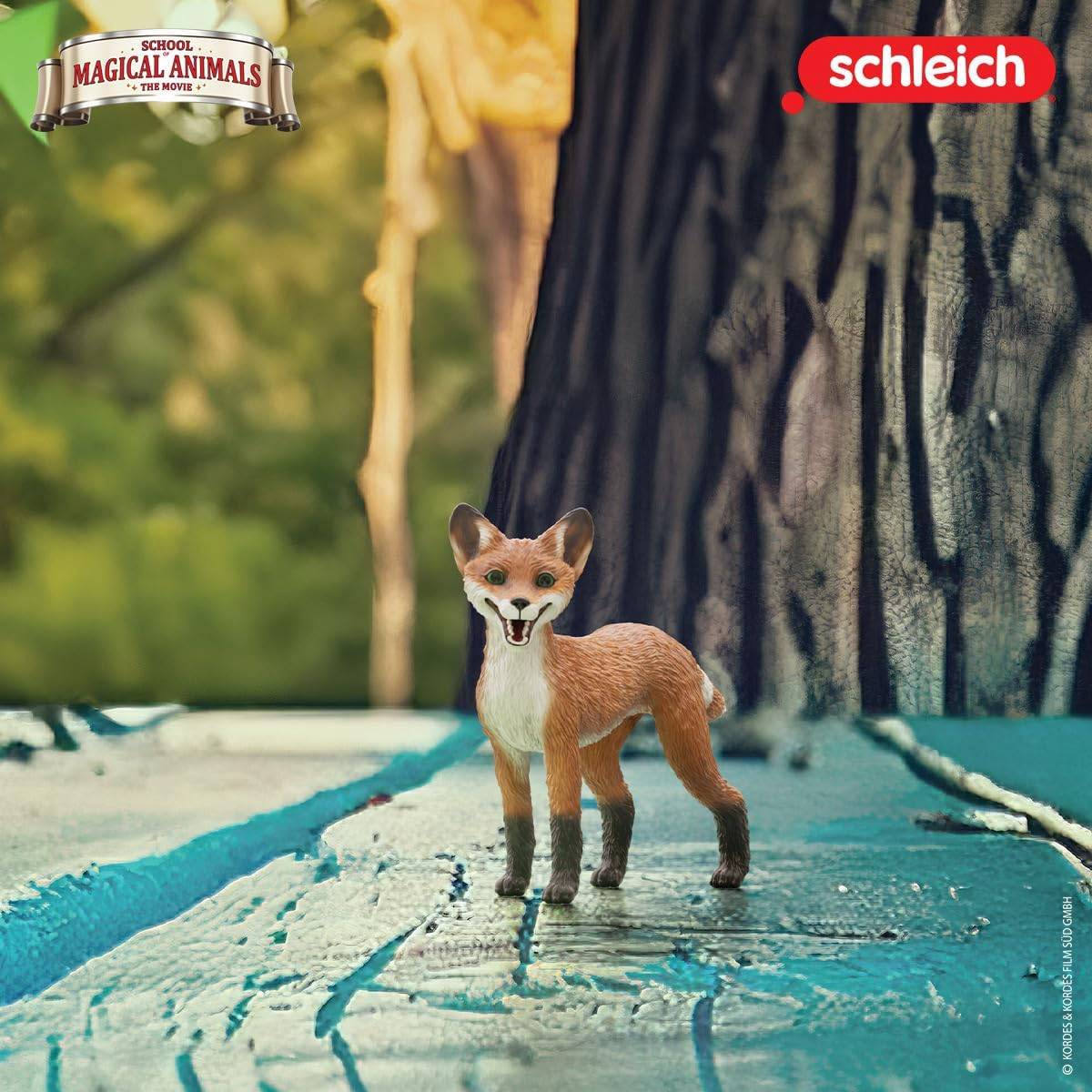 Eine kleine Spielzeug-Fuchsfigur steht auf einer strukturierten blauen Oberfläche mit einem Baumstamm im Hintergrund. Die Logos 'School of Magical Animals' und 'Schleich' sind sichtbar.
