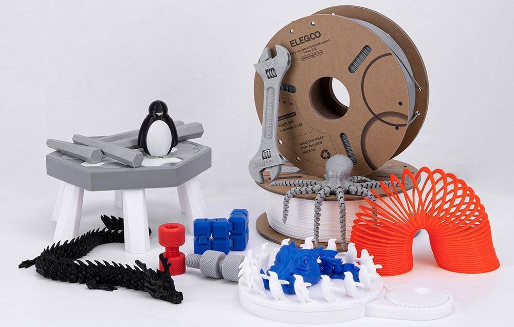 3D-gedruckte Objekte wie ein Schraubenschlüssel, Zahnräder und eine Pinguin-Figur, die um Filamentspulen herum ausgestellt sind.