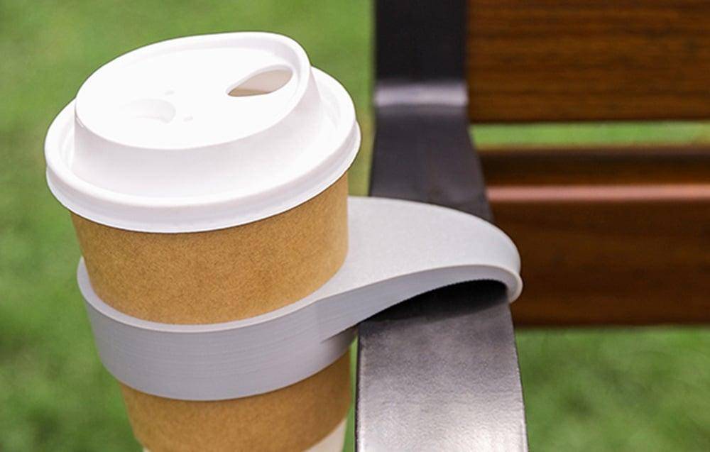 Eine Kaffeetasse mit Deckel steht in einem kreisförmigen Halter, der an der Armlehne einer Bank auf einer Rasenfläche befestigt ist.