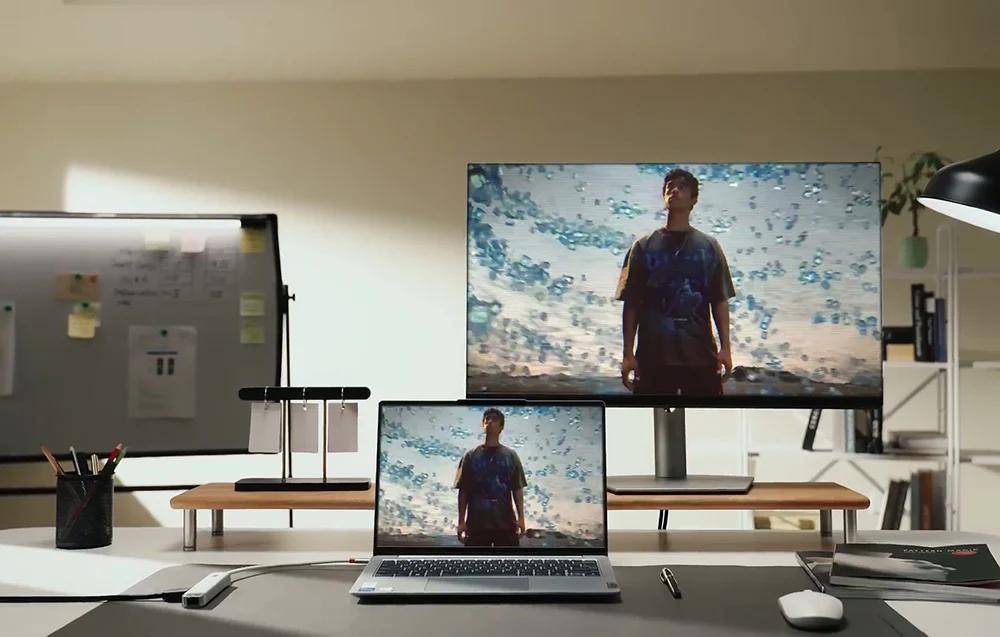 Ein Laptop und ein Monitor zeigen dasselbe Bild einer stehenden Person mit einer malerischen Kulisse. Büromaterial und eine Whiteboard sind zu sehen.