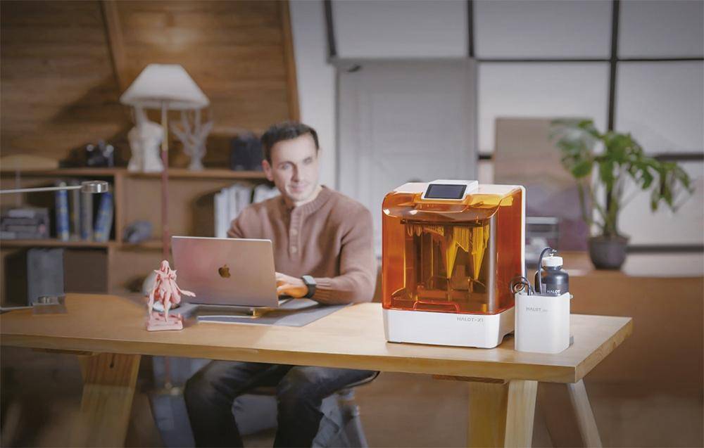 Ein Mann, der an einem Laptop an einem Schreibtisch arbeitet, neben einem orangefarbenen 3D-Drucker mit einem kleinen Modell und einer Lampe im Raum.