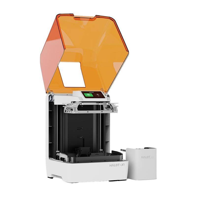 Ein 3D-Drucker mit einem offenen, orangefarbenen Deckel, beschriftet mit 'HALOT-XT'. Das Gerät ist neben einem kleinen externen Bauteil positioniert.
