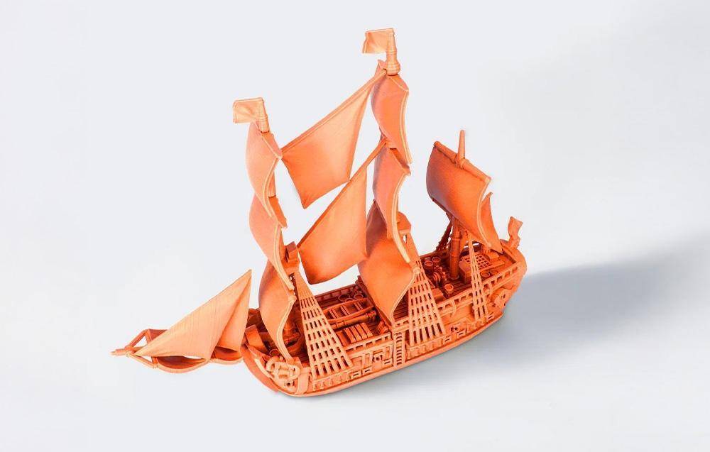 Ein kompliziertes orangefarbenes 3D-gedrucktes Modell eines Segelschiffs mit detaillierten Masten, Segeln und Rumpf, das auf einem weißen Hintergrund präsentiert wird.